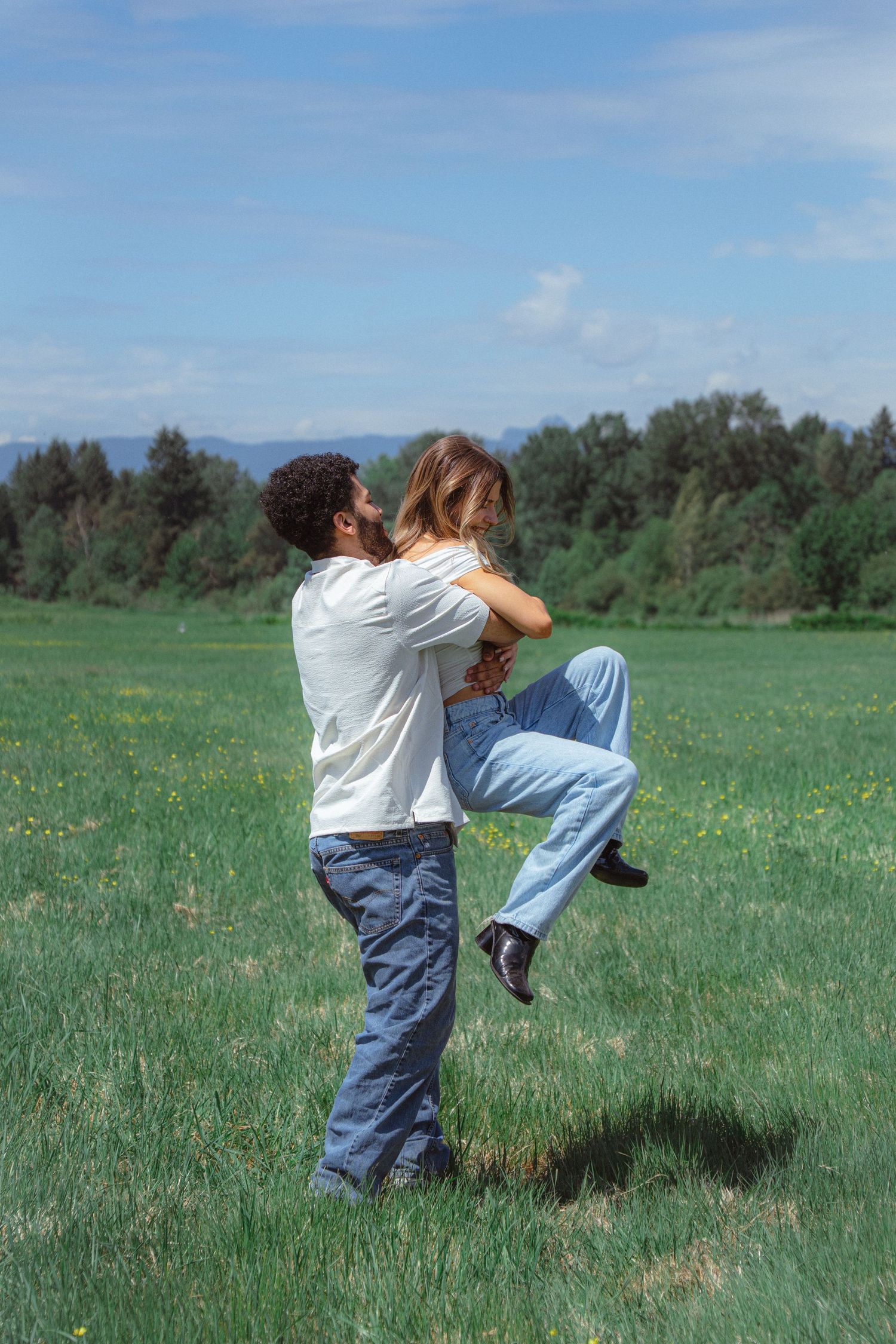 A man lifts a woman joyfully in a green meadow under a blue sky with trees in the background.