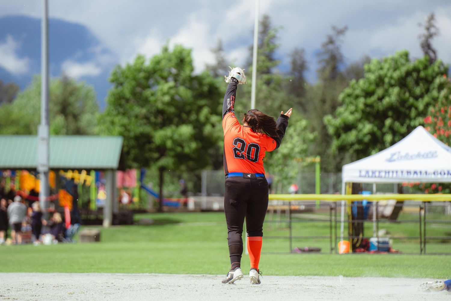 Softball player number 20 in orange uniform raises arms celebrating on outdoor field with trees behind.