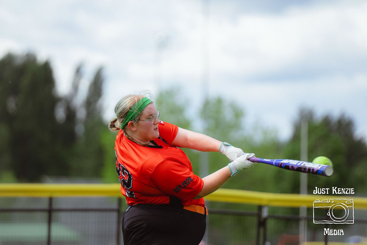 Young female softball player in orange jersey swinging bat during a competitive outdoor game.