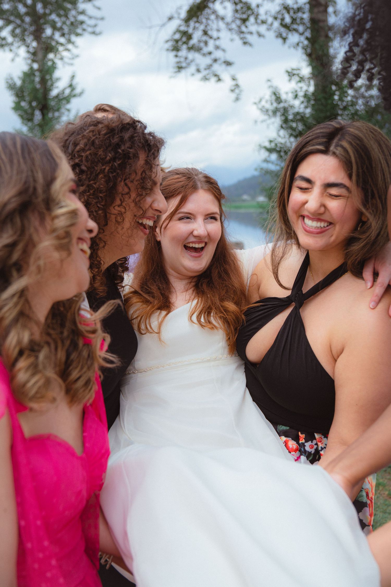 Bride laughs joyfully surrounded by smiling bridesmaids at outdoor wedding celebration.