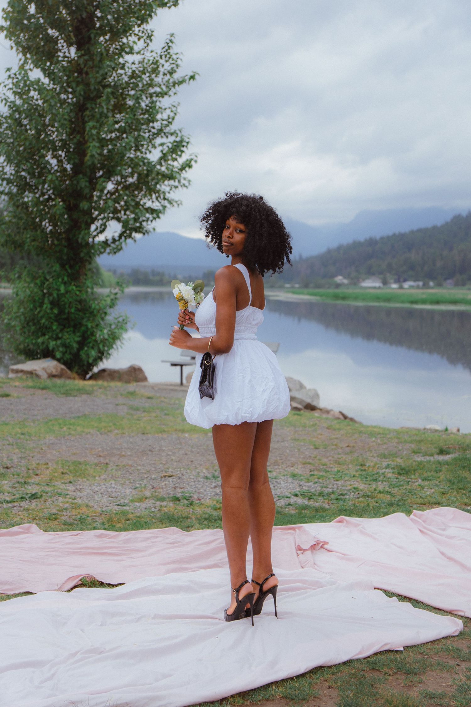 Black woman in white bubble dress holding flowers standing on pink fabric by a mountain lake.