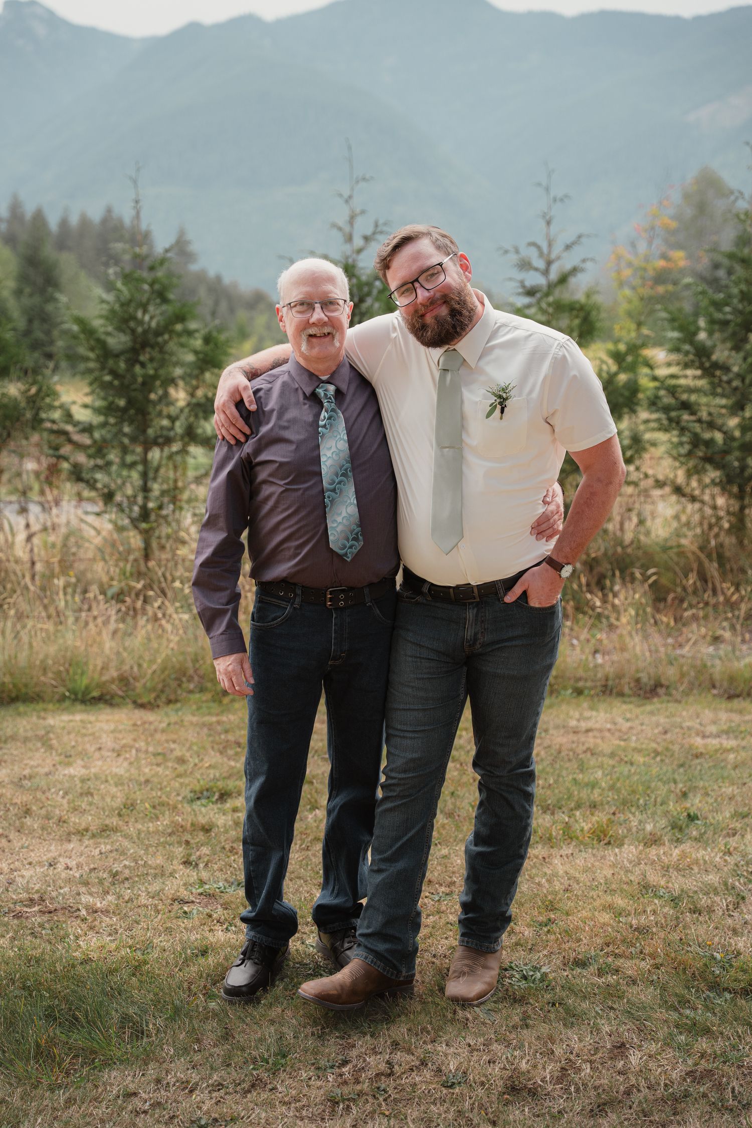 Two men smiling together outdoors in ties with scenic mountain background at a wedding.