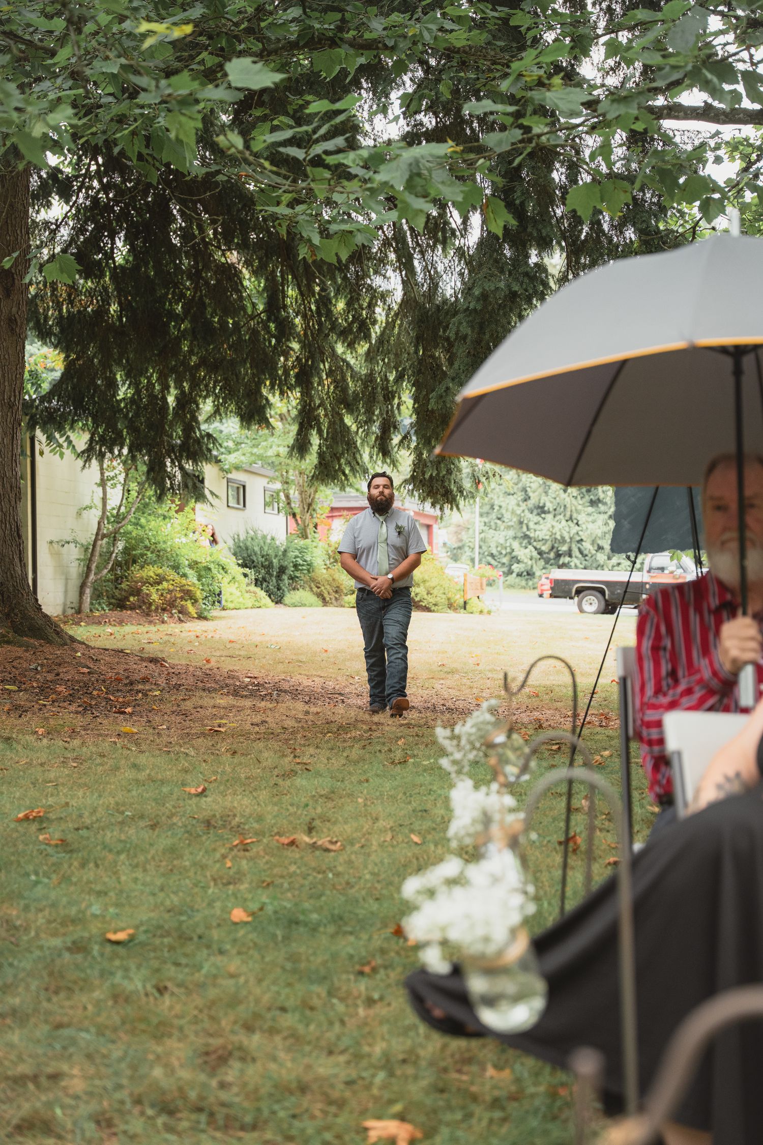 Bearded man walks toward wedding guests seated under umbrella at outdoor garden ceremony surrounded by trees.