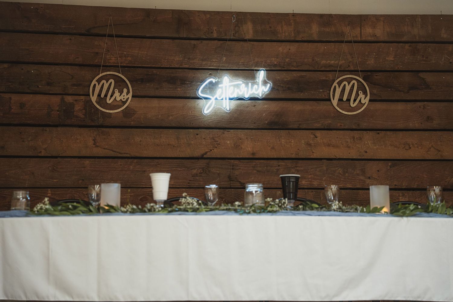 Rustic wooden wedding head table with neon sign, Mrs and Mr signs, candles, and greenery garland centerpiece.