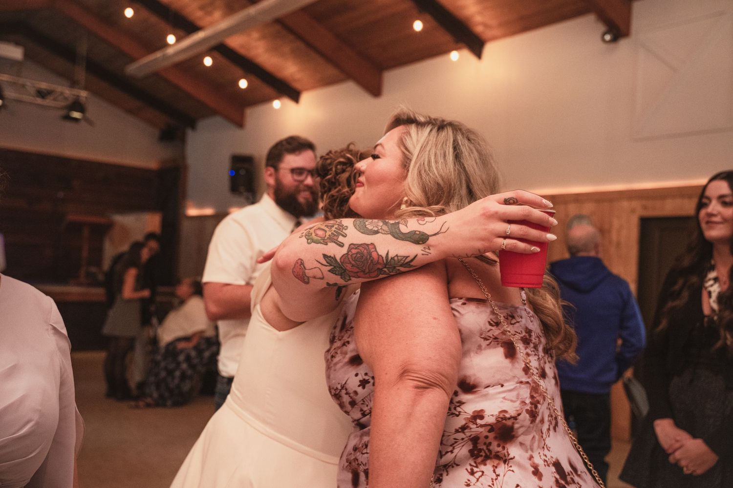 Bride with tattoos sharing a warm hug with a guest in floral dress at wedding reception.