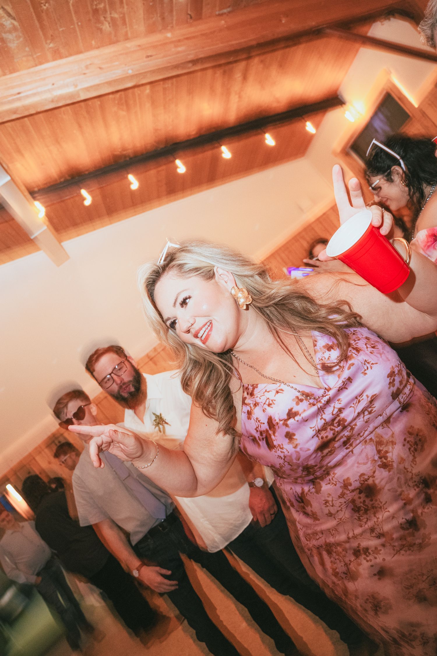 Blonde woman in floral dress dancing at a party, holding a drink, with guests and string lights in the background.