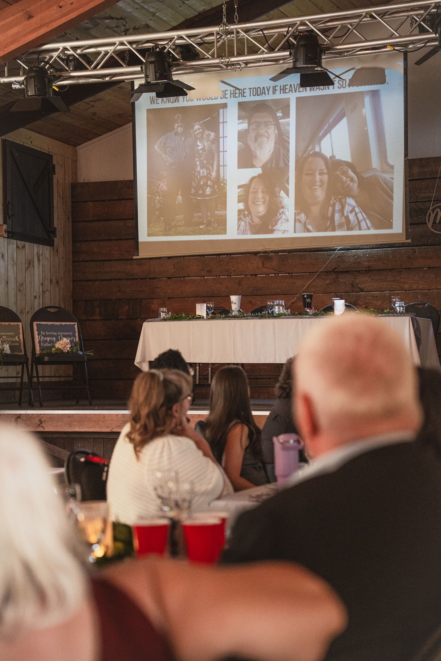 Wedding guests watch a touching memorial photo slideshow projected on screen in a rustic barn reception hall.