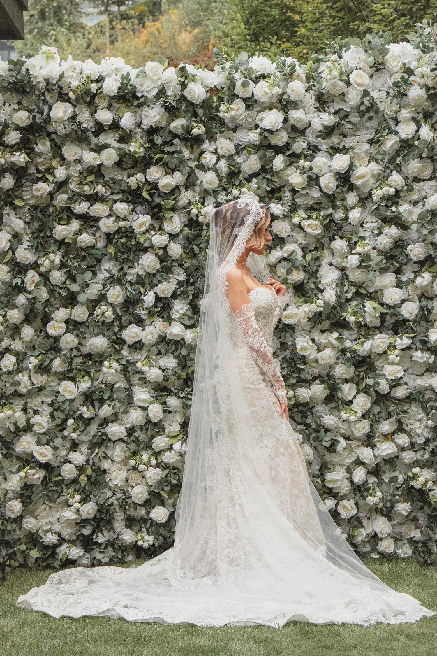Bride in lace wedding dress with cathedral veil stands before lush white rose flower wall backdrop.