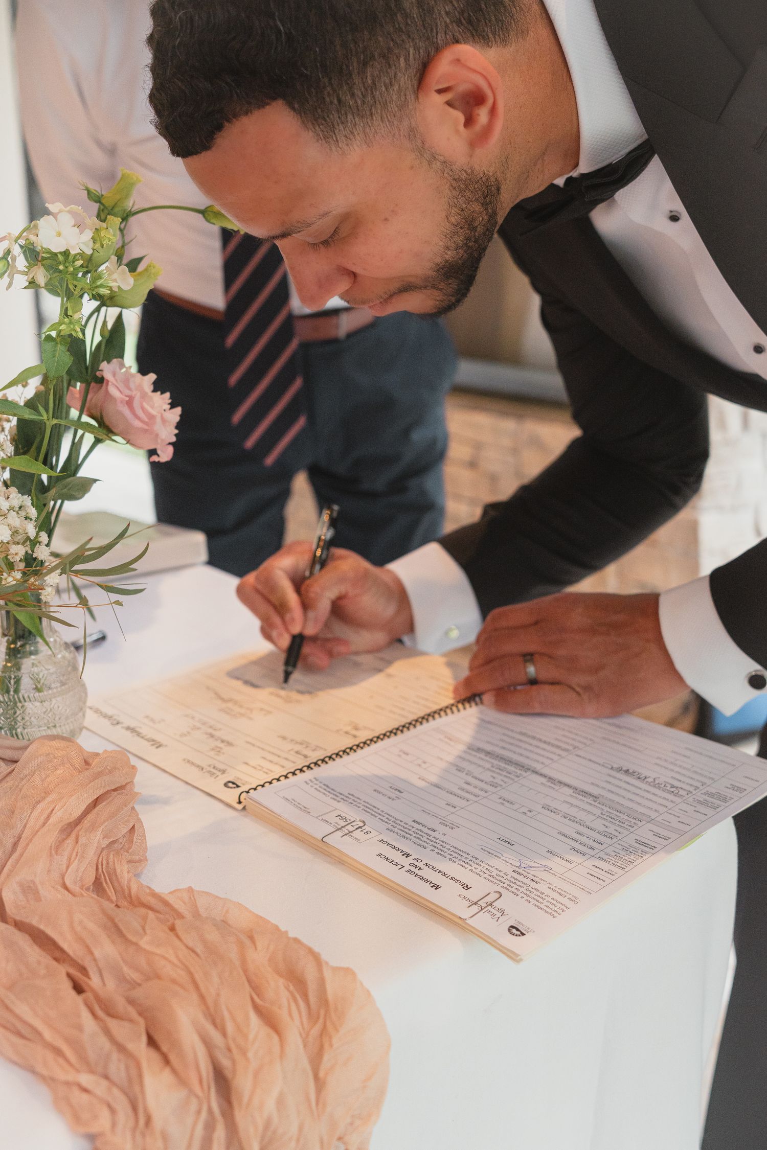 Groom in tuxedo signing marriage certificate at decorated table with flowers.