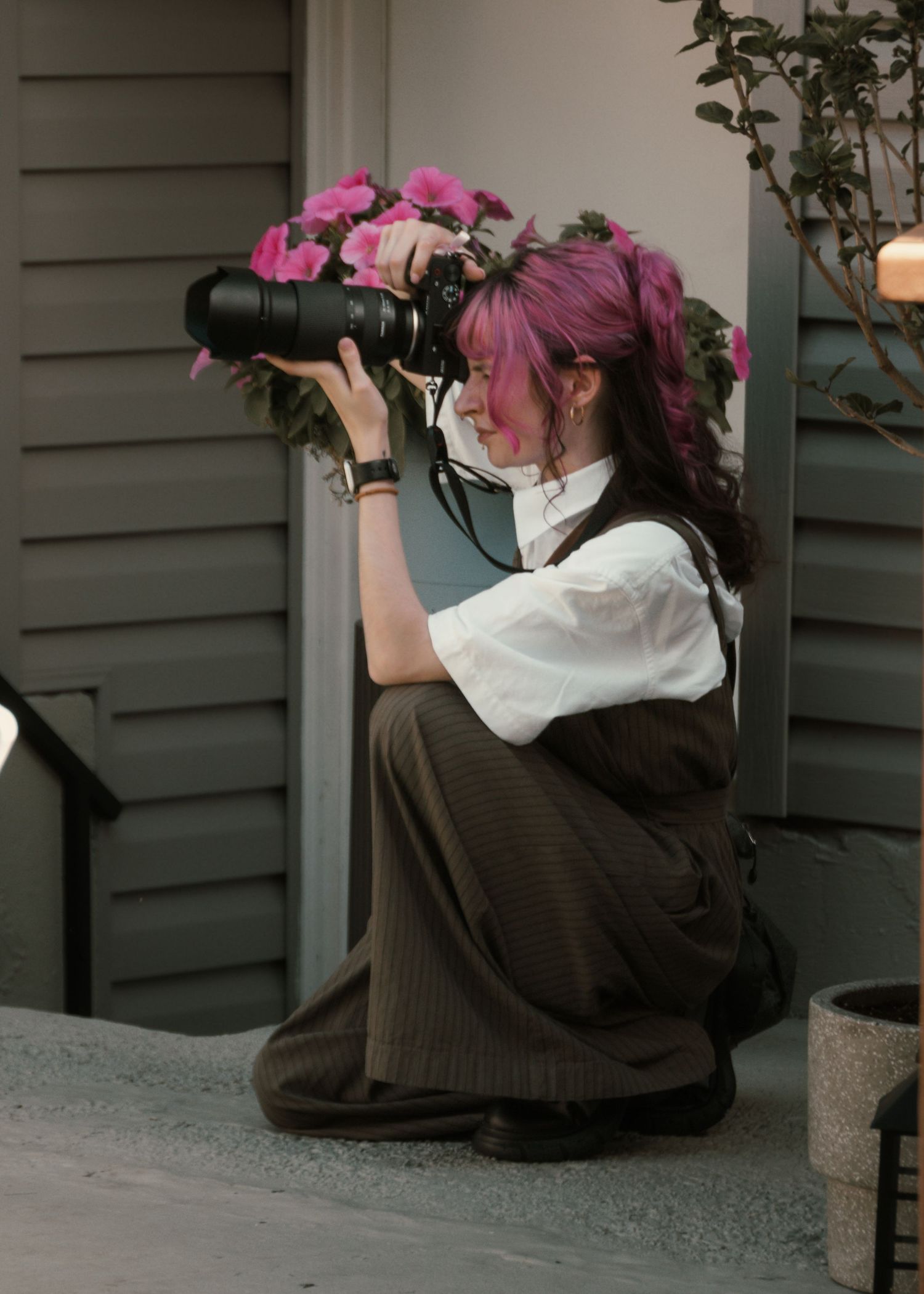 Woman with pink hair crouching while using a professional camera, wearing a white blouse and dark vest outdoors.