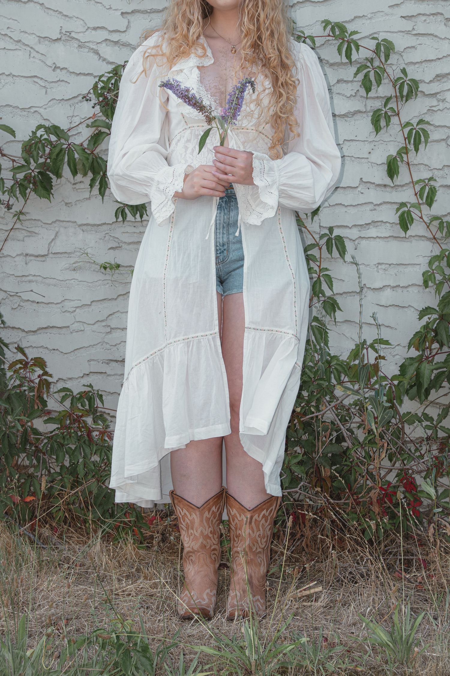 Woman in white boho duster dress, denim shorts and cowboy boots holding purple lavender bouquet outdoors.