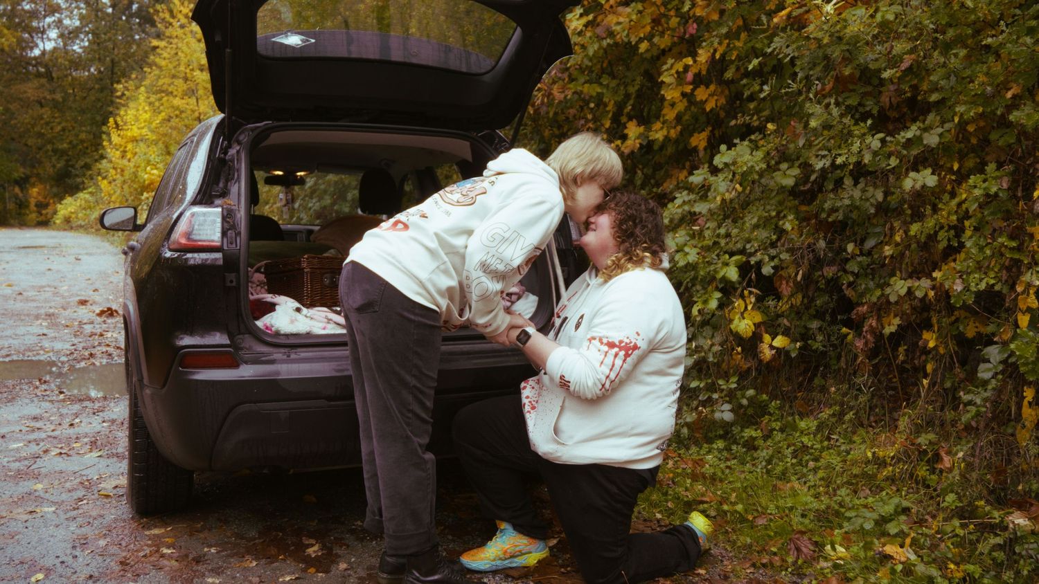 Two people in white hoodies lean into the trunk of a dark SUV surrounded by autumn foliage.
