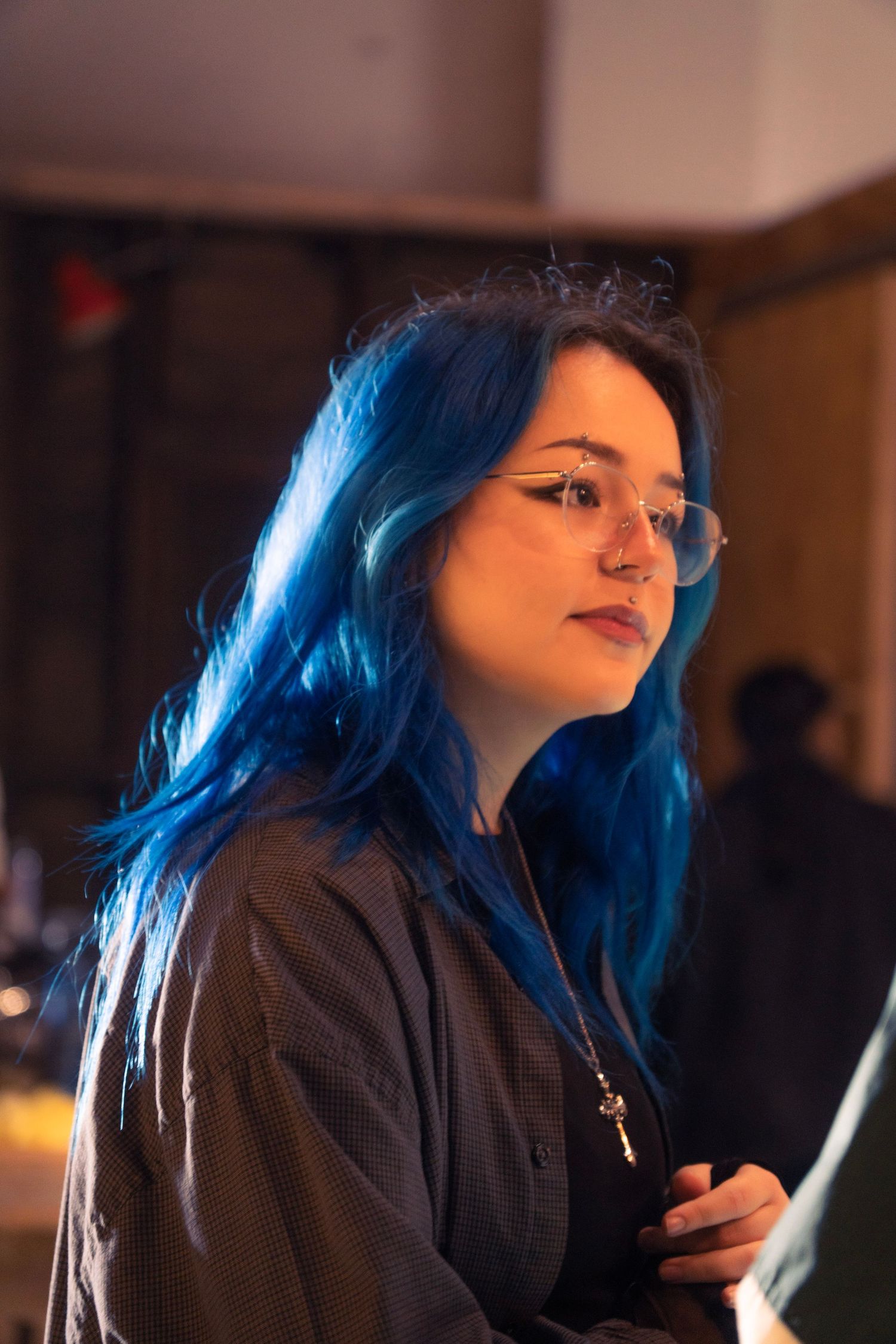 Young woman with vibrant blue hair and glasses looking thoughtfully in a warmly lit room.