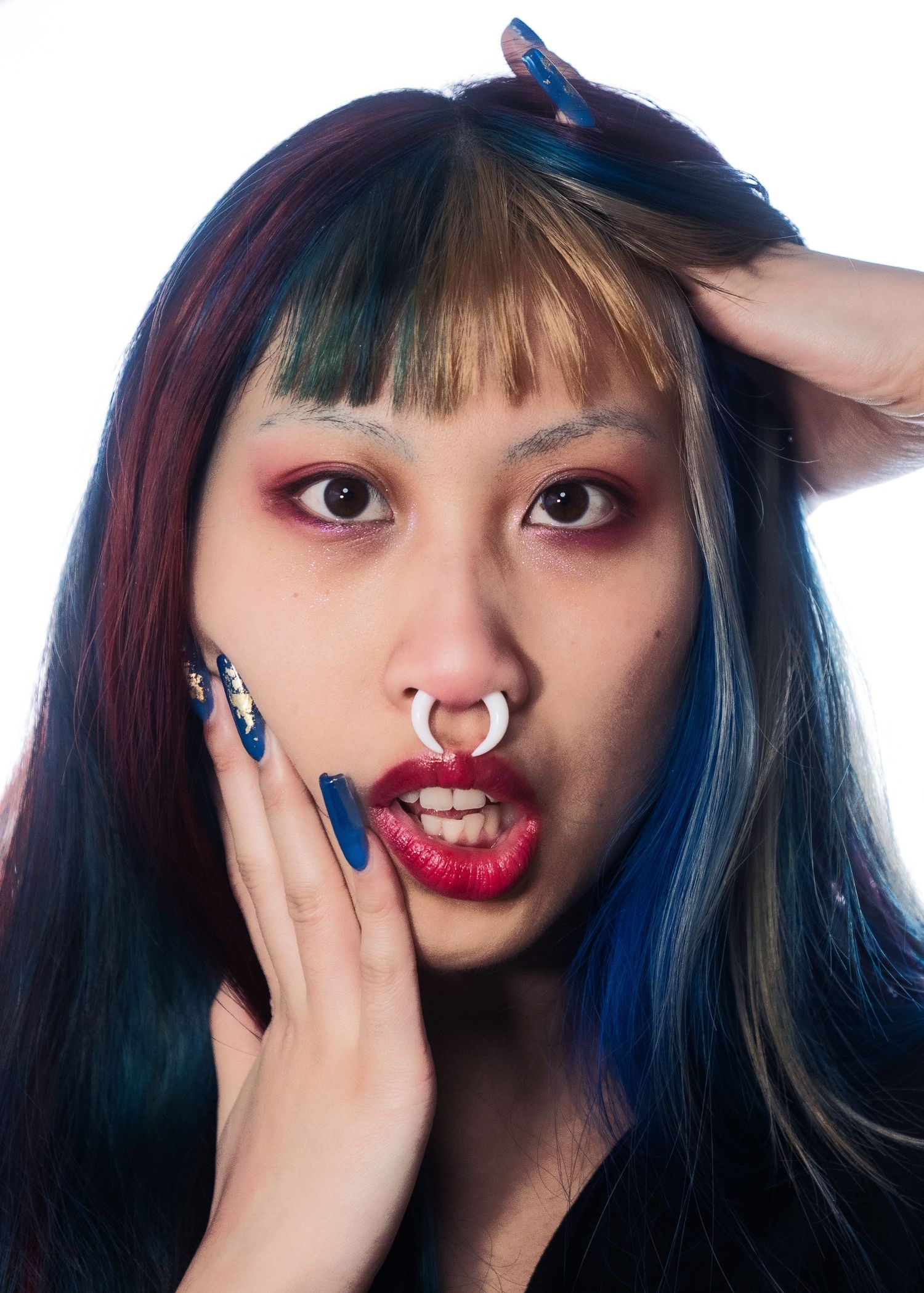 Young woman with colorful blue and red hair, septum piercing, bold makeup, and blue nail art.
