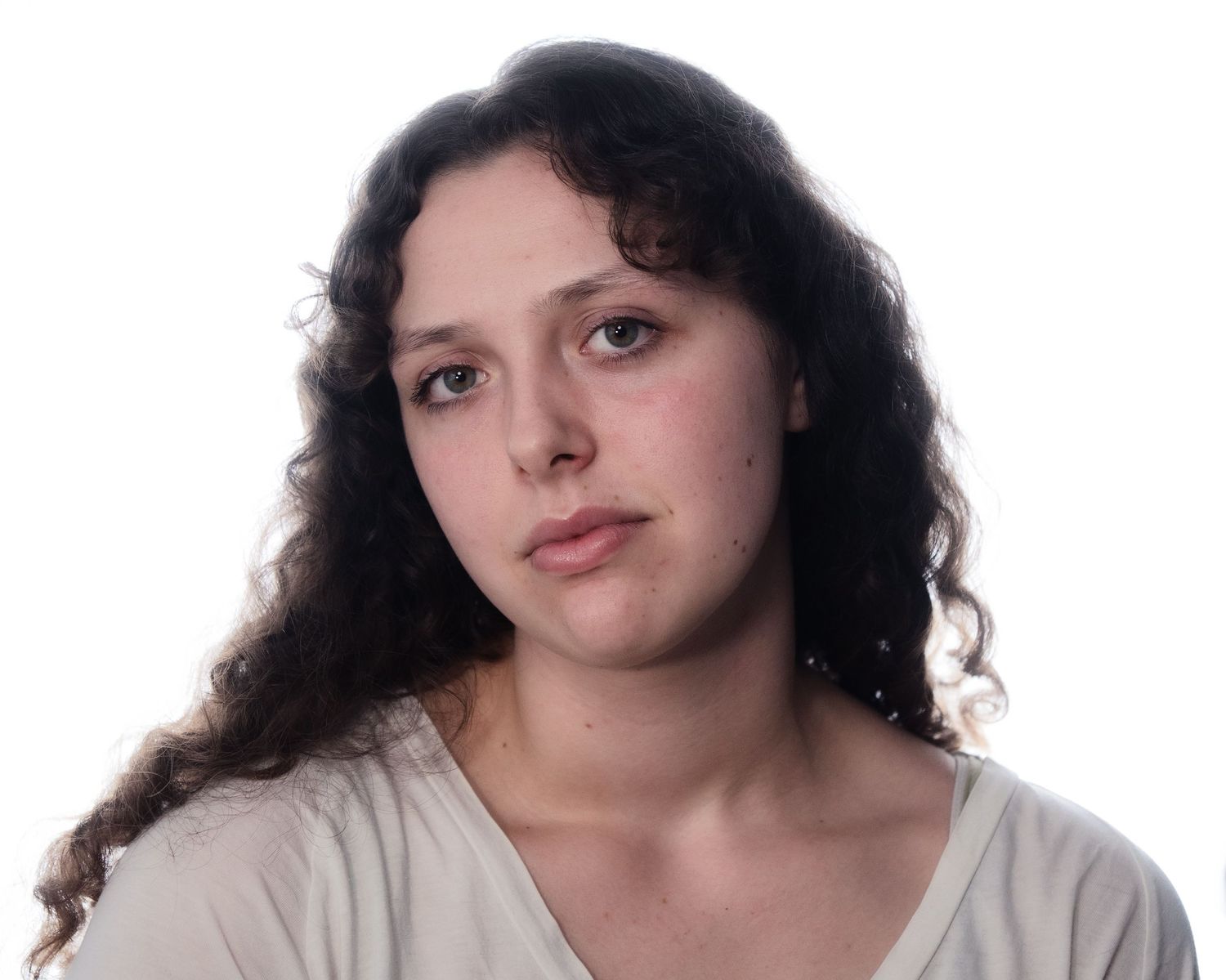 Young woman with long curly dark hair in neutral expression wearing a white top against white background.
