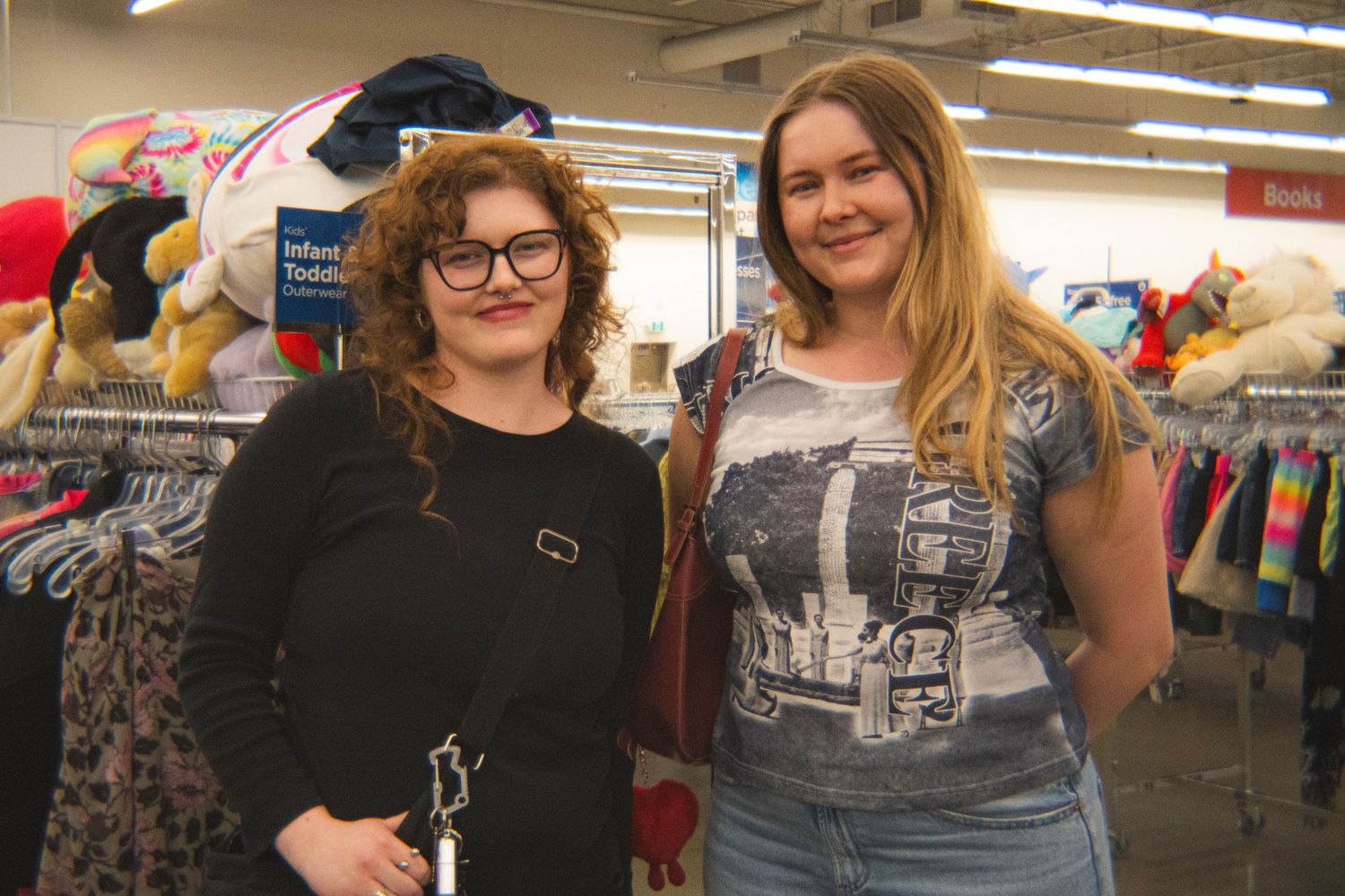 Two female friends posing together while browsing a thrift store clothing section.