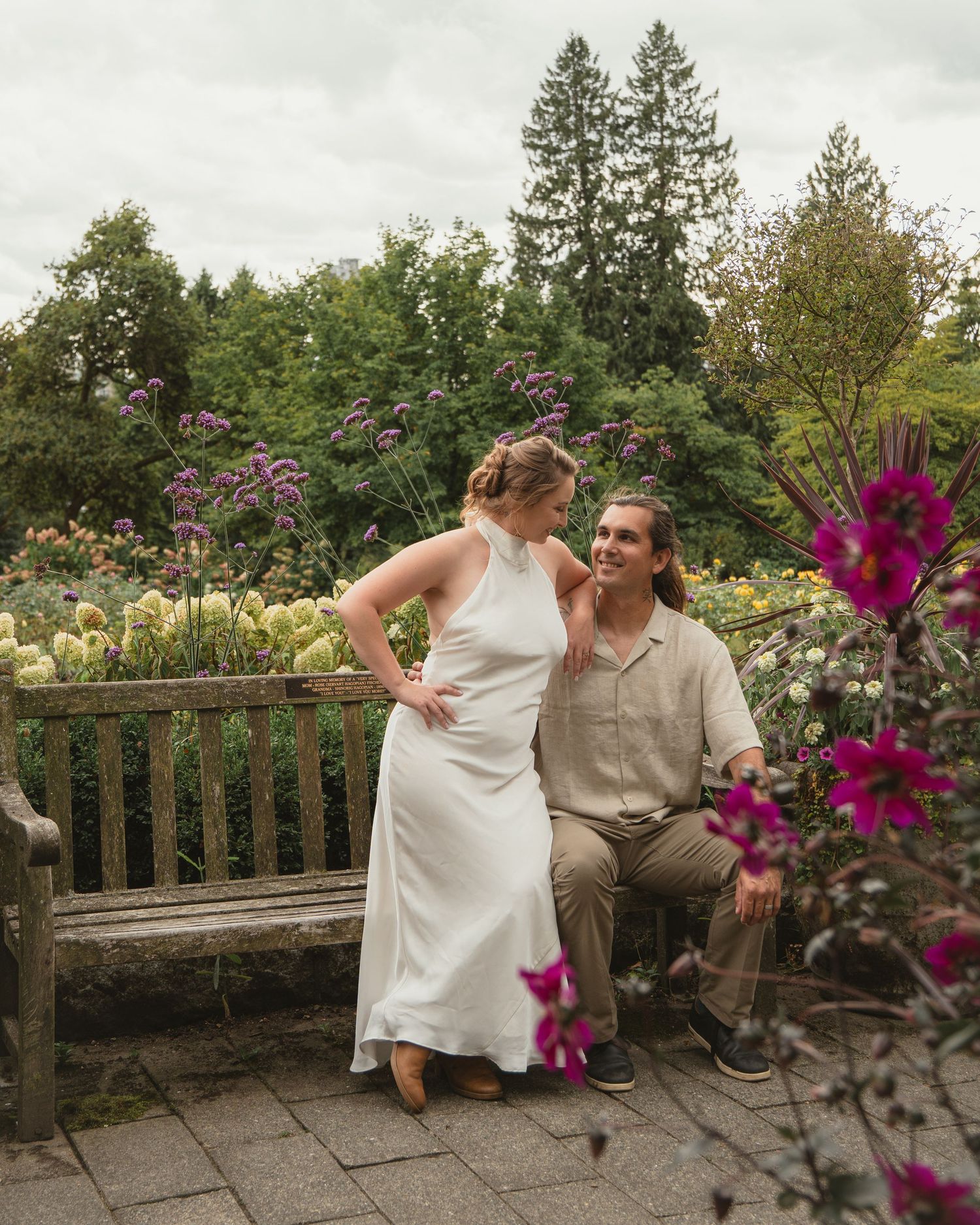 Bride in white halter dress and groom in beige shirt pose by a wooden bench in a blooming garden.