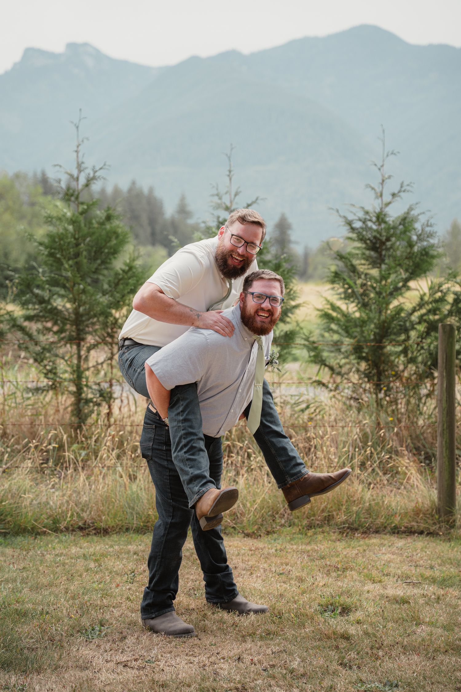 Two men laughing as one gives the other a piggyback ride outdoors with mountains behind.