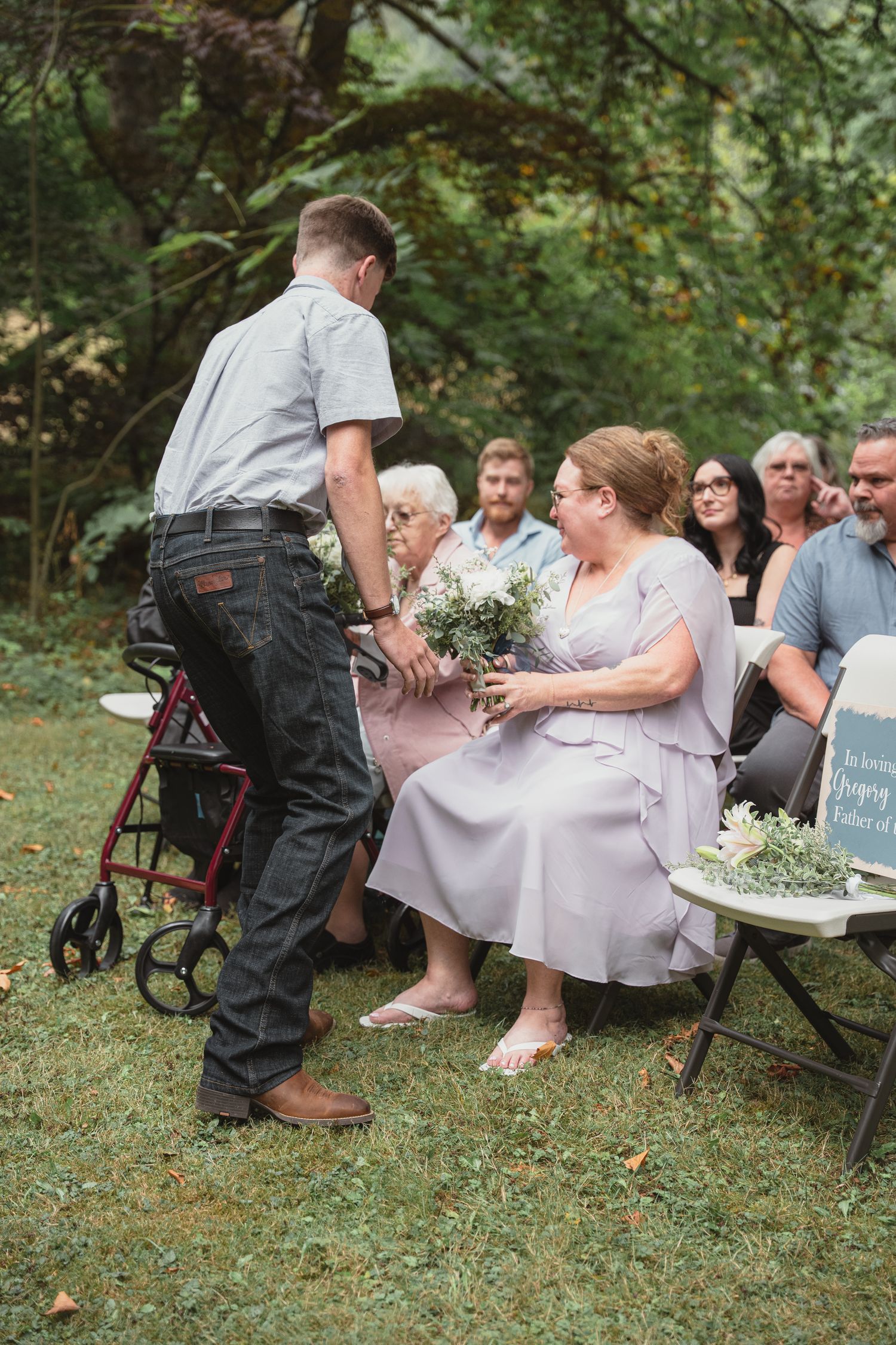 Groomsman hands bouquet to seated woman in lavender dress at outdoor wedding ceremony.