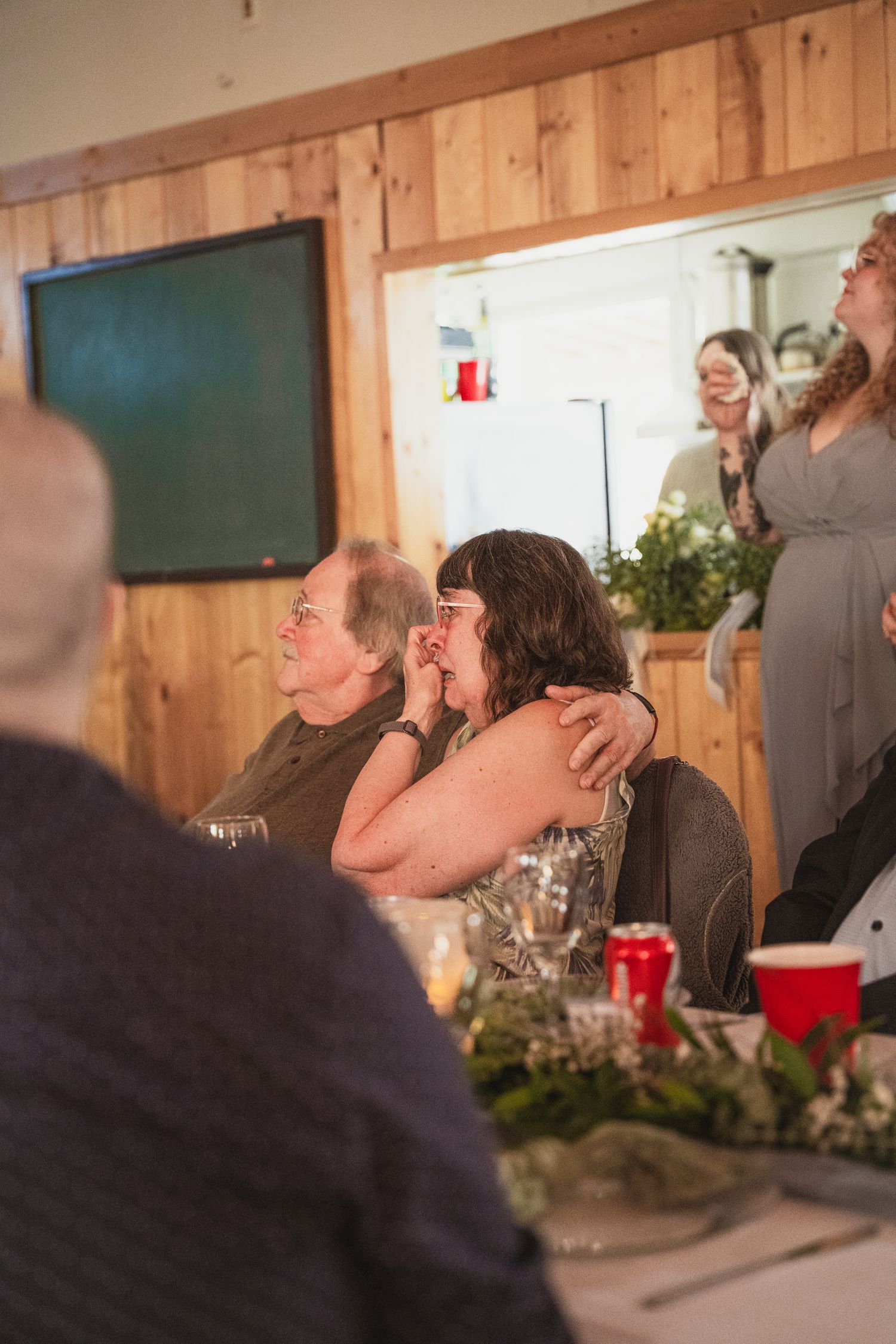 Wedding guests sharing an emotional embrace while watching a presentation at a rustic reception venue.