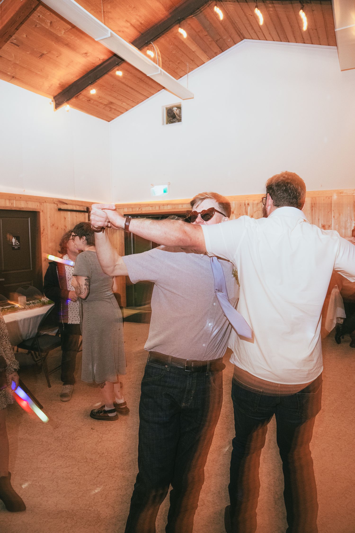 Two men dancing together at an indoor wedding reception venue with wood ceiling and glowing light sticks.