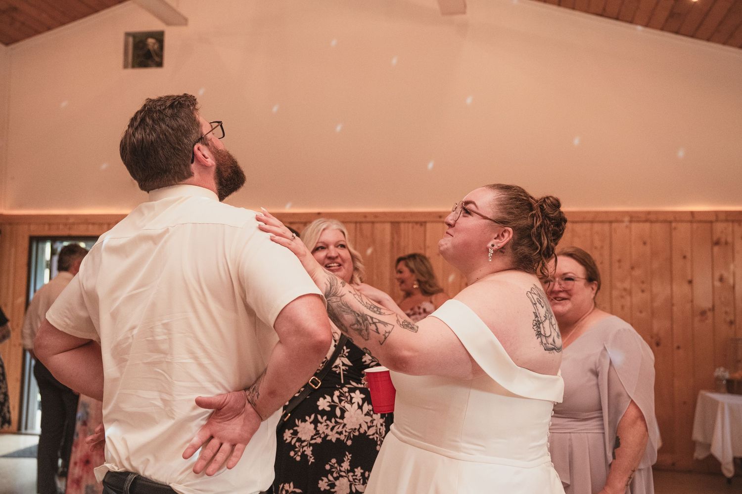 Bride and groom laugh and dance joyfully with guests during lively rustic barn wedding reception.