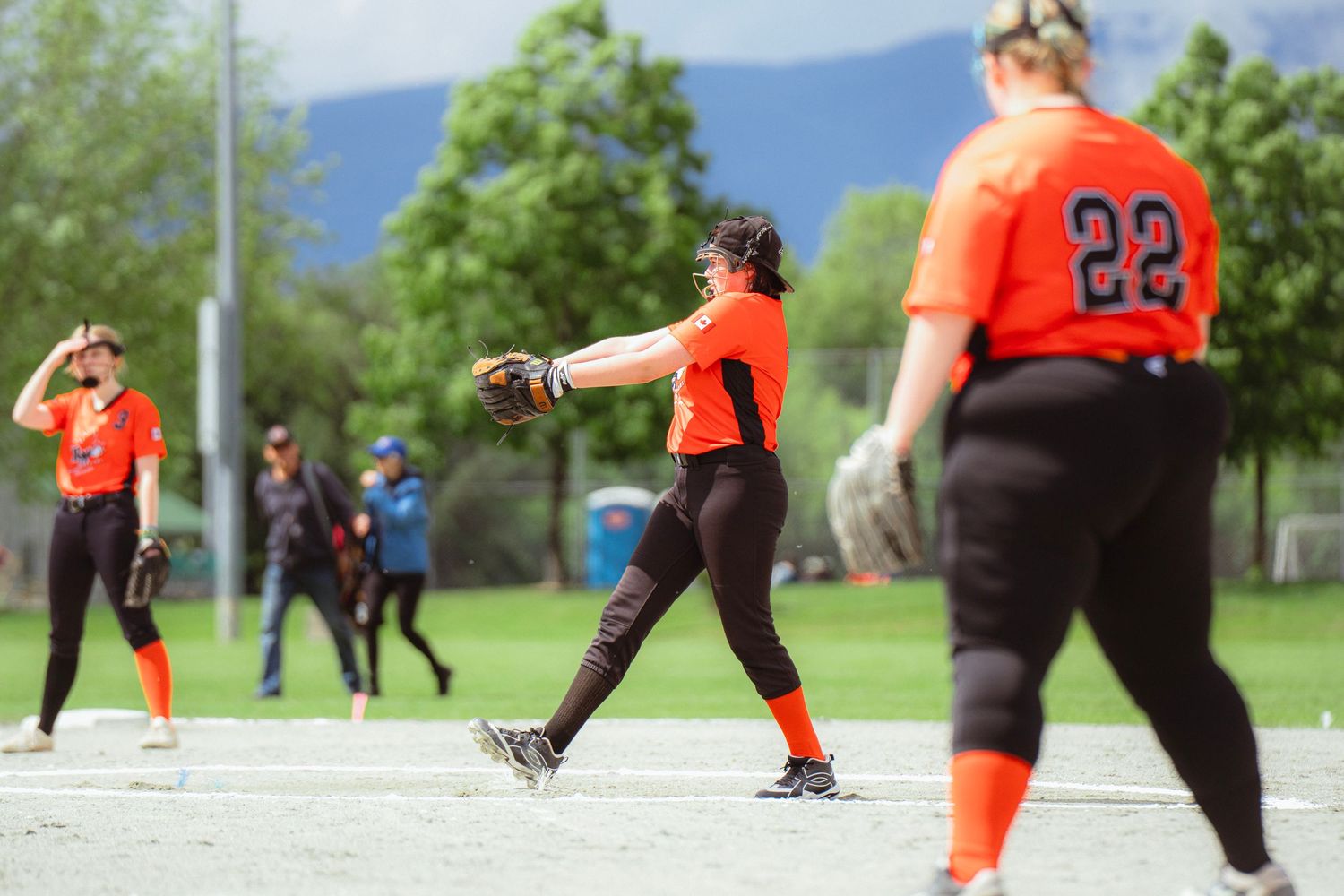 Softball player in orange jersey throwing ball during outdoor game with teammate number 22 in foreground.