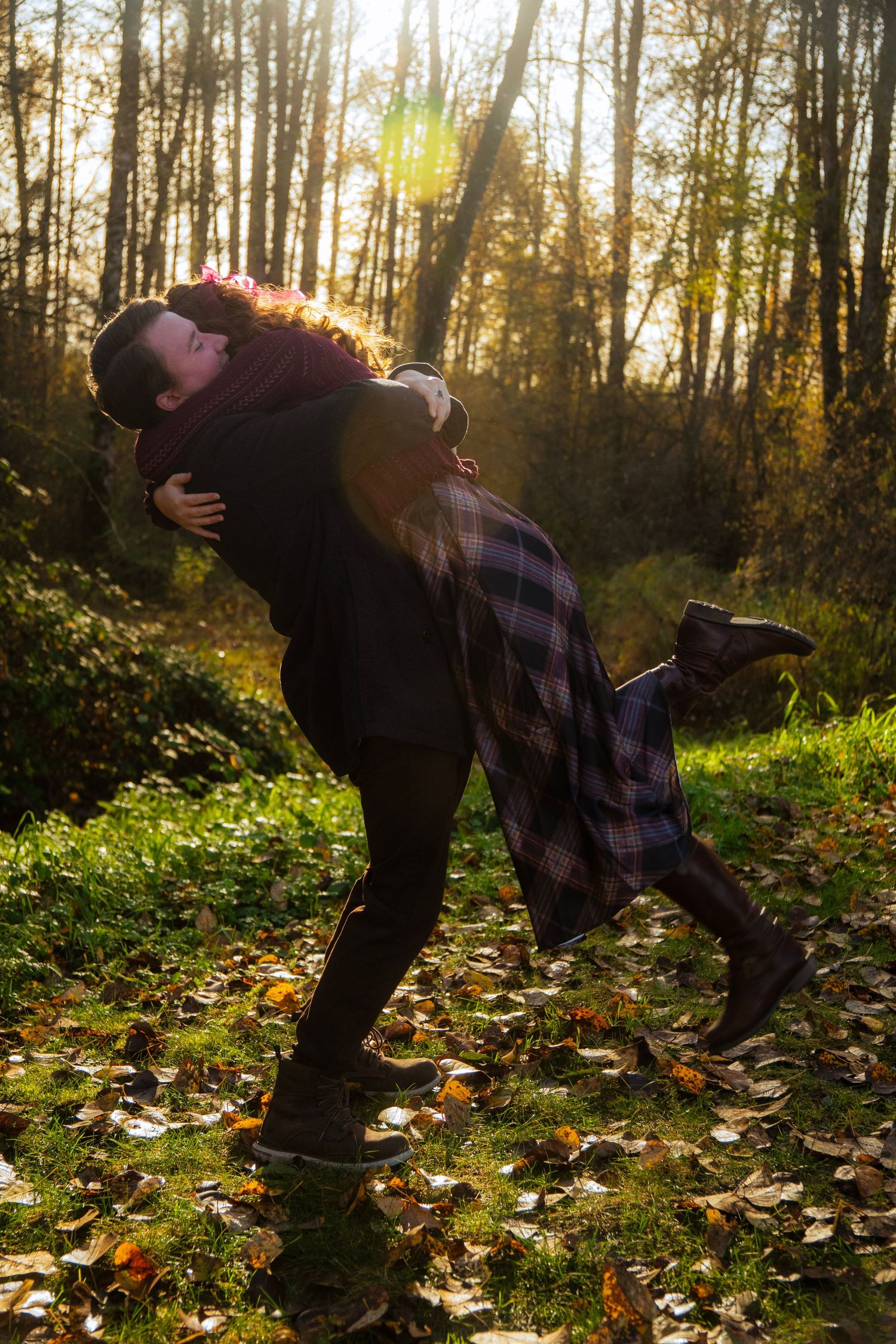 Man lifting woman in a joyful embrace in a sunlit autumn forest with fallen leaves on the ground.