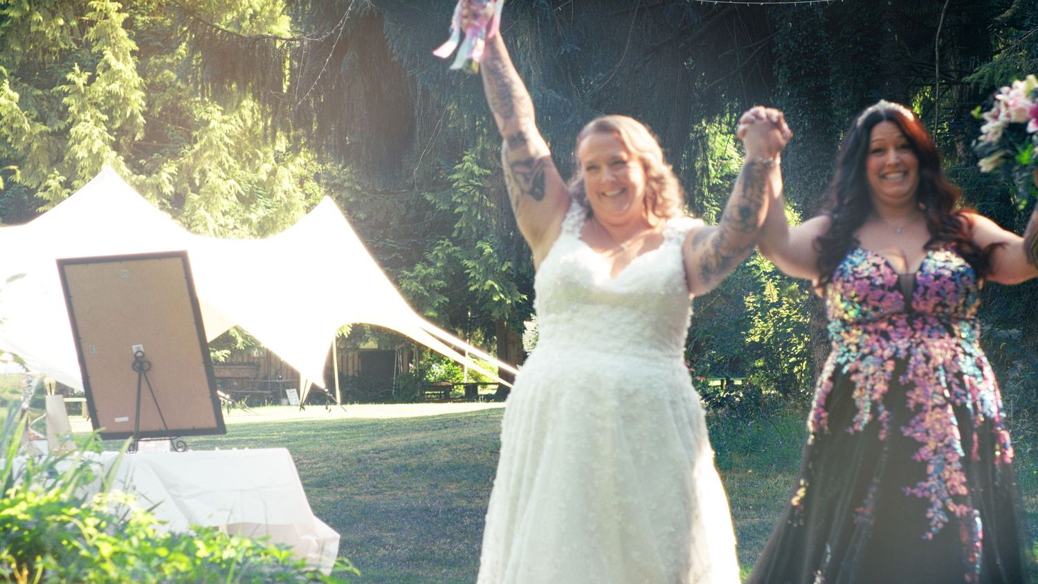 Bride in white gown celebrating with raised arms at an outdoor garden wedding ceremony with a tent backdrop.