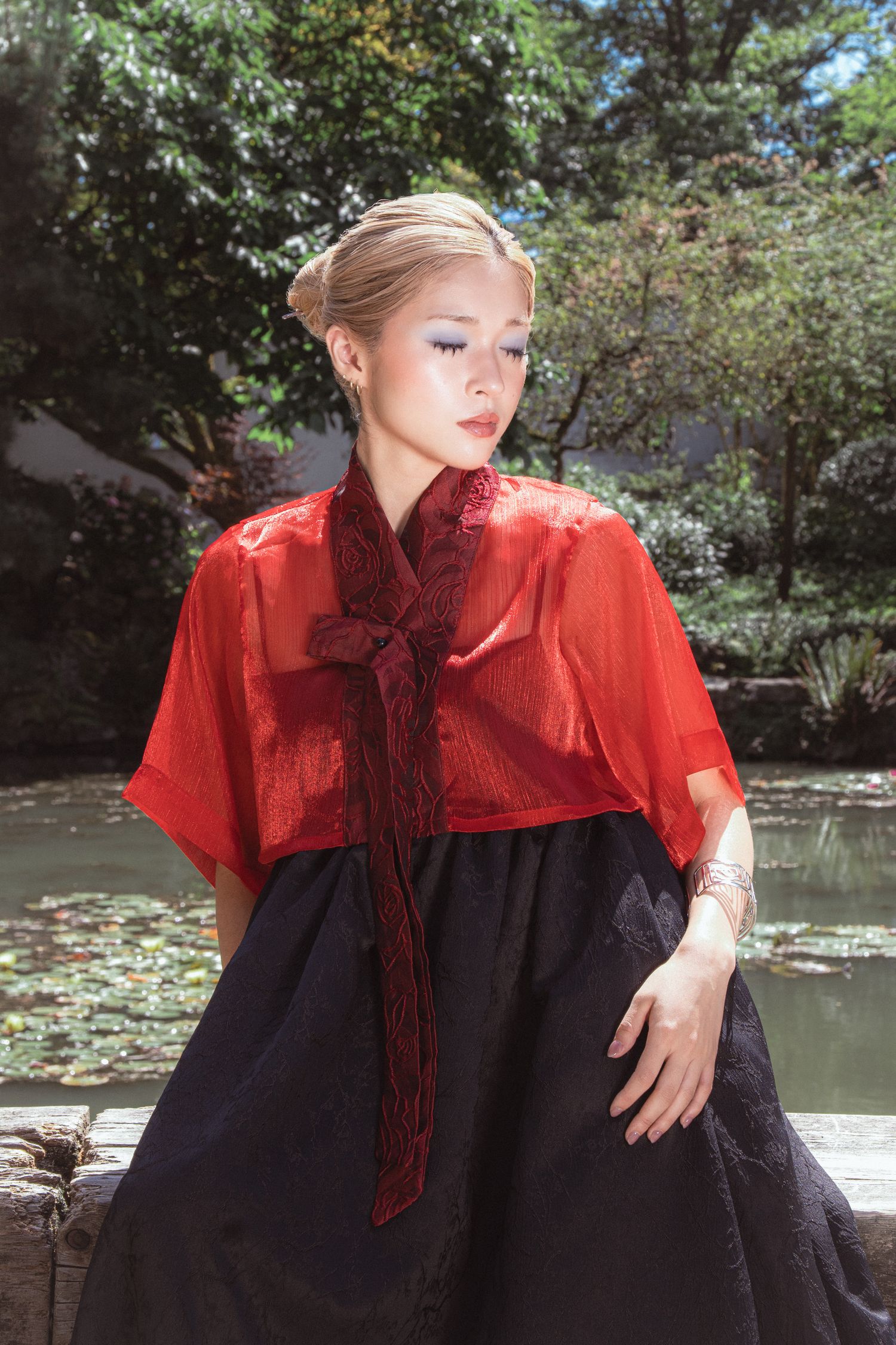 Young woman in red sheer top and black skirt poses gracefully outdoors near a serene pond with lily pads.