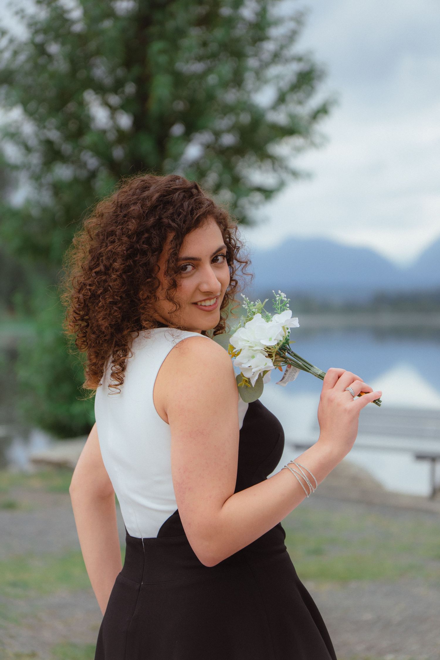 Curly-haired woman in black and white dress holding white flowers smiling by a mountain lake.