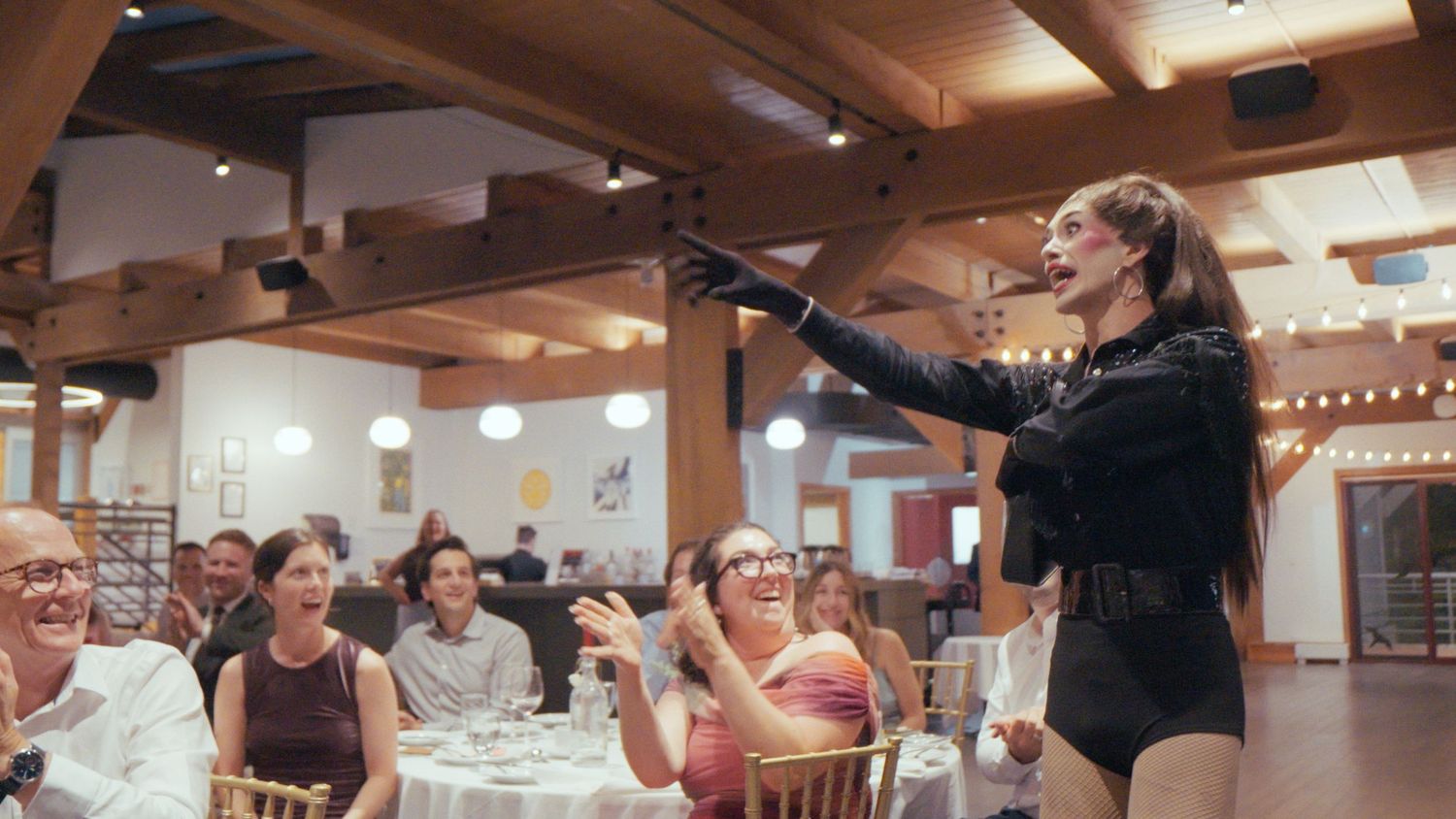 An entertainer in a black bodysuit performs energetically for laughing guests at a rustic wedding venue.