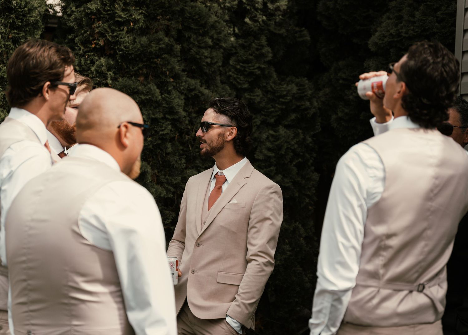 Groom in tan suit with sunglasses stands with groomsmen in white shirts outdoors near green trees.