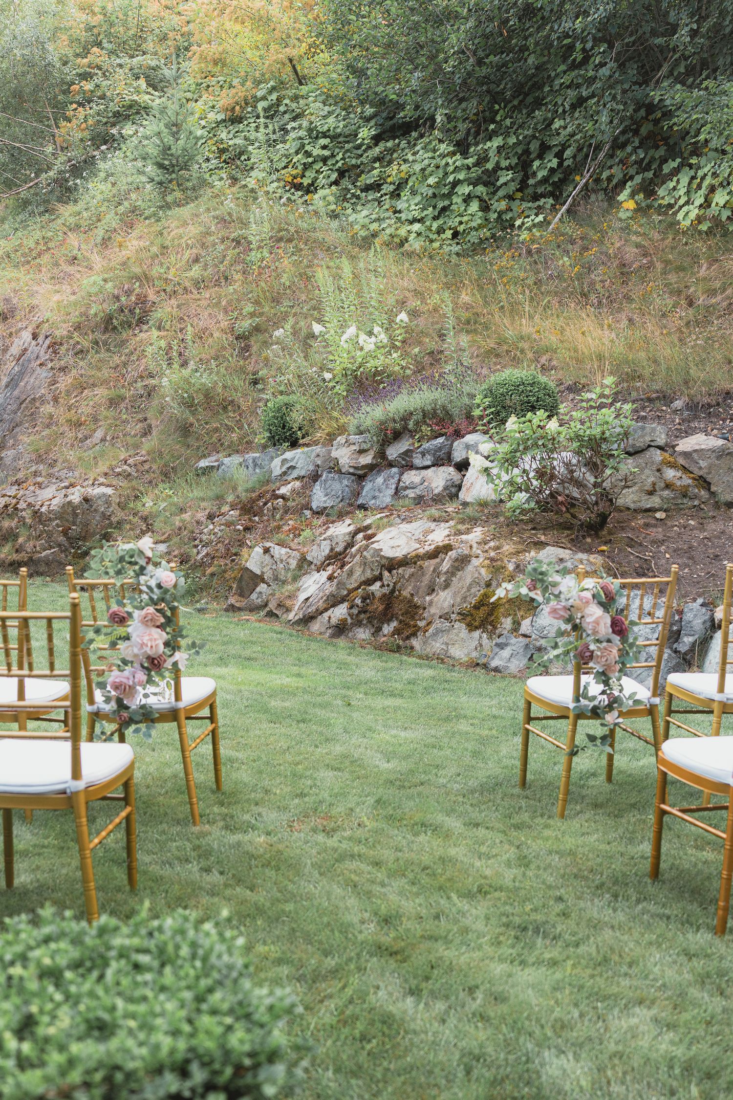 Outdoor wedding ceremony setup with gold chiavari chairs adorned with floral arrangements on a lush green lawn.
