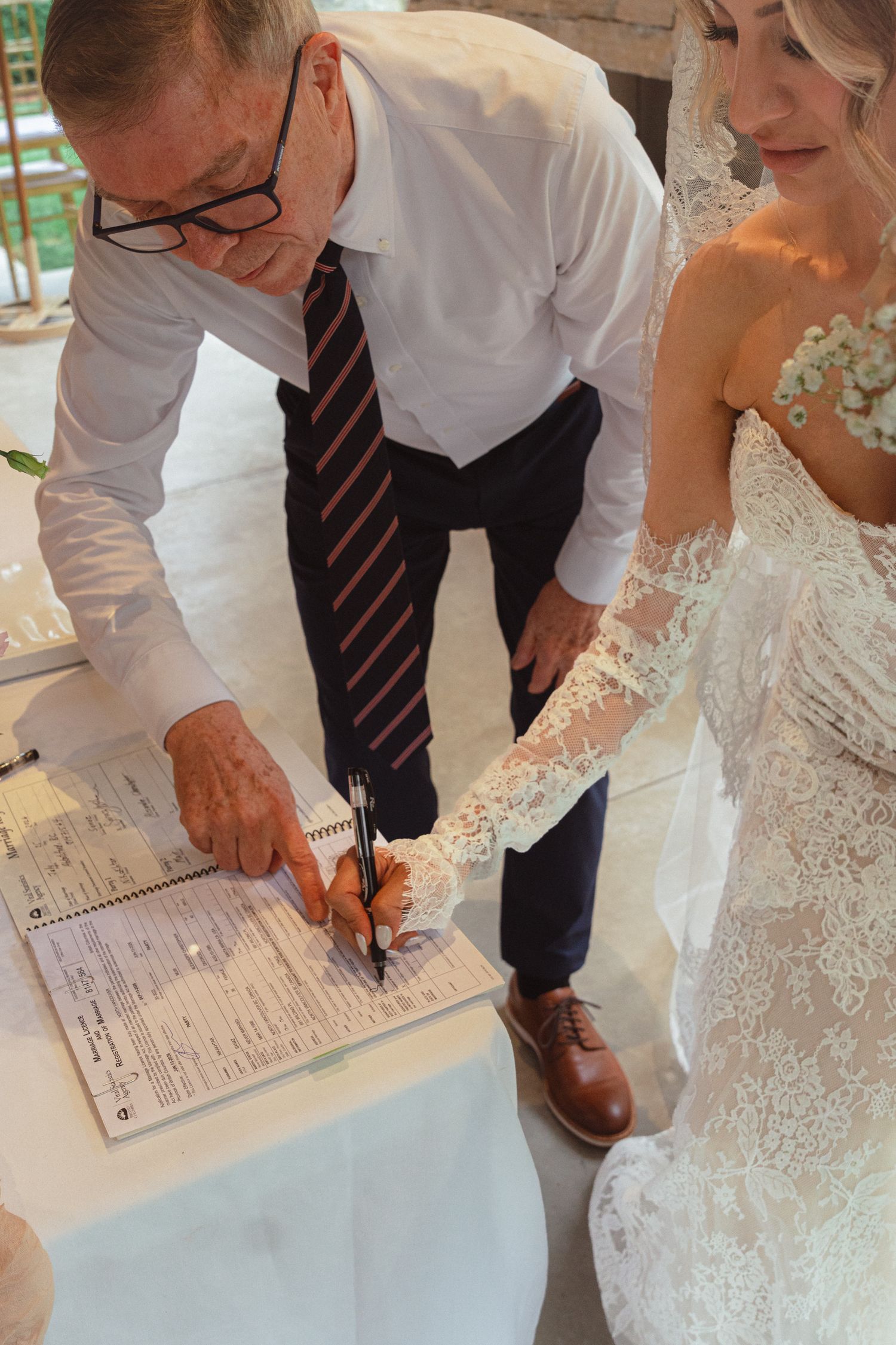Older man signing wedding certificate beside bride in lace wedding dress during ceremony.