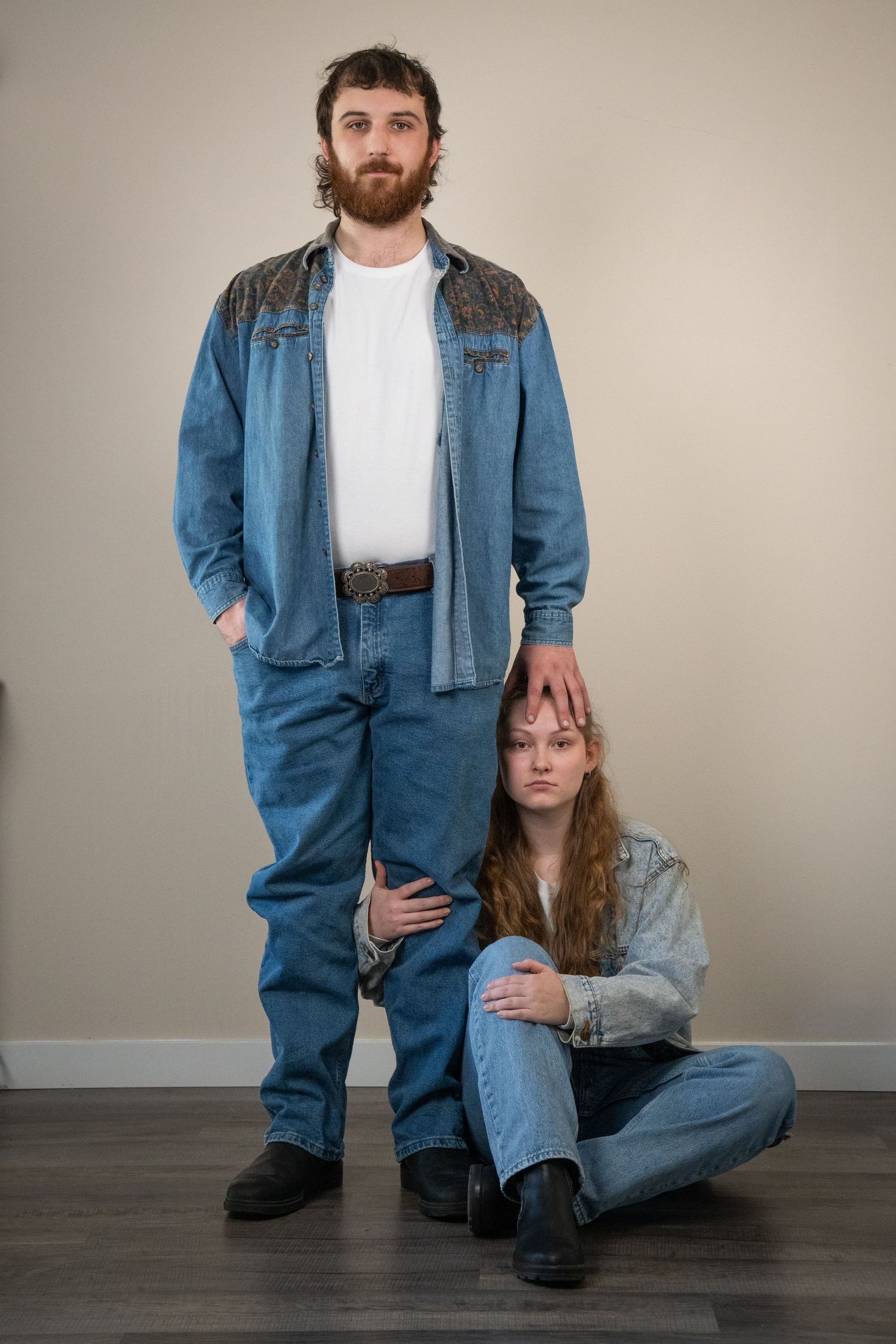 A bearded man stands with hand on seated womans head, both wearing matching denim outfits in a studio setting.