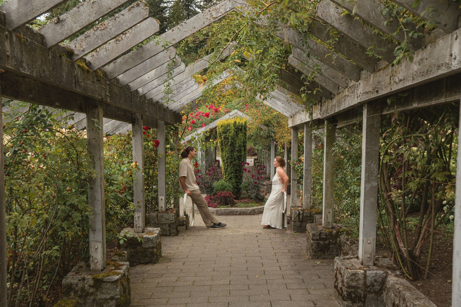 Couple in wedding attire walking through a lush garden pergola with stone columns and colorful foliage.