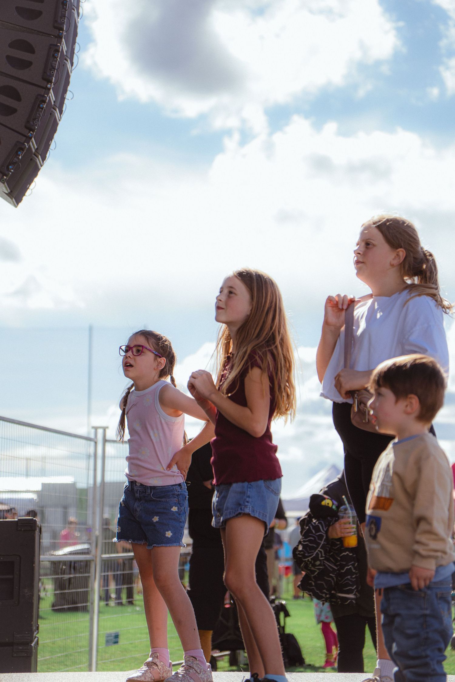 Four children watching outdoor music festival performance on sunny day with cloudy blue sky.