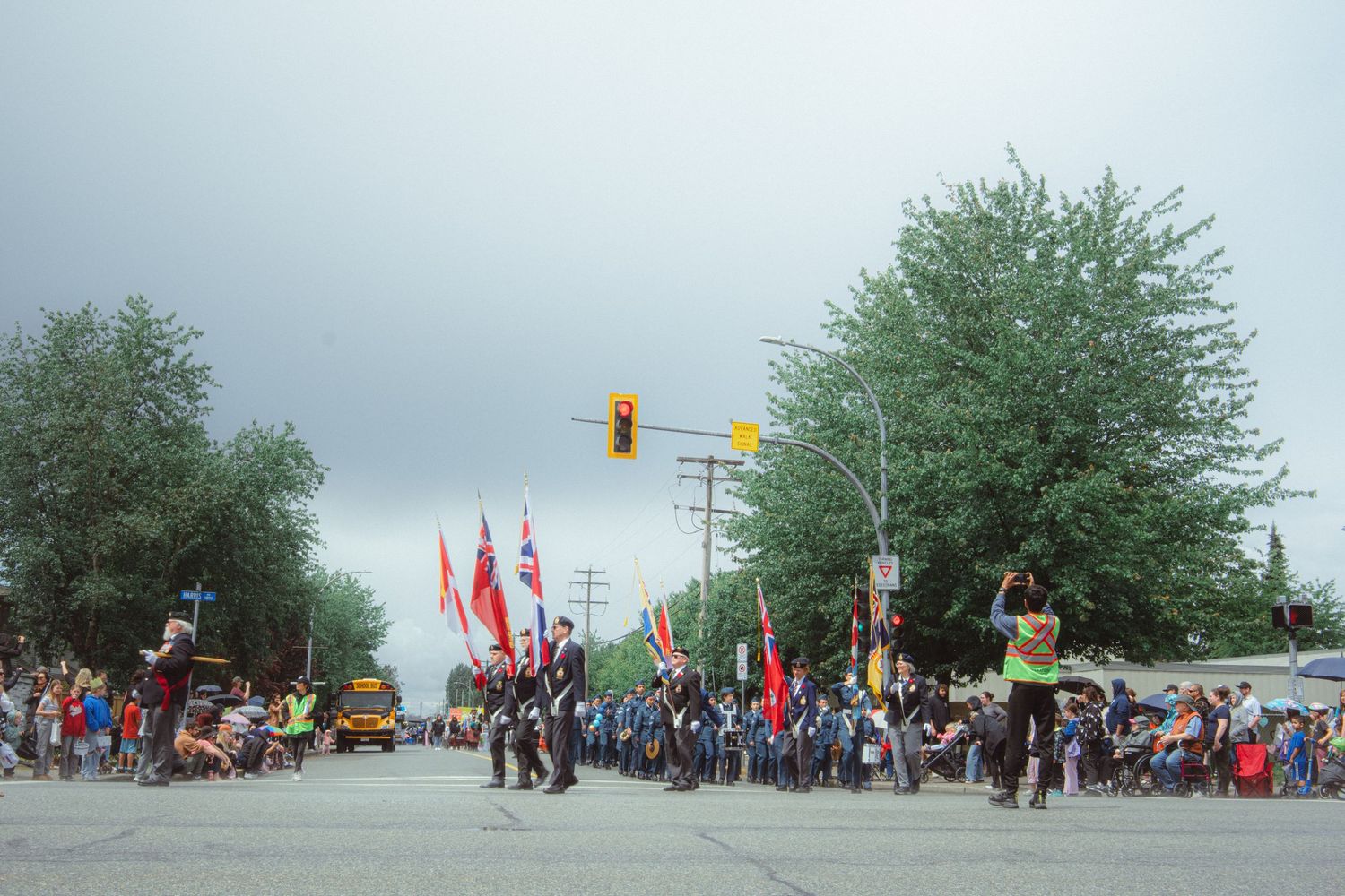 Marching band members carry flags and parade down a street intersection under overcast skies.