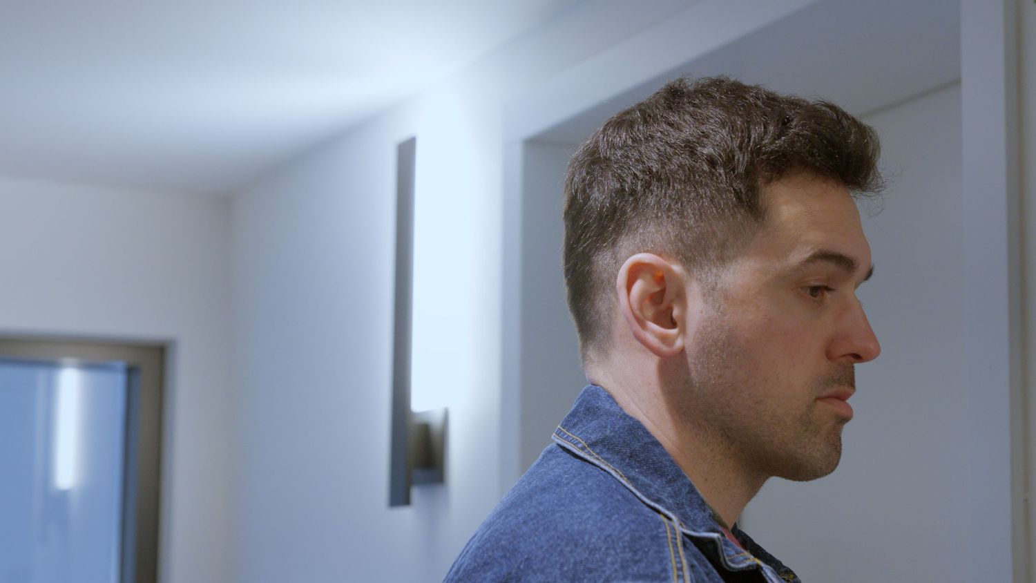 Side profile of a young man with short hair in a denim jacket standing in a bright white room.