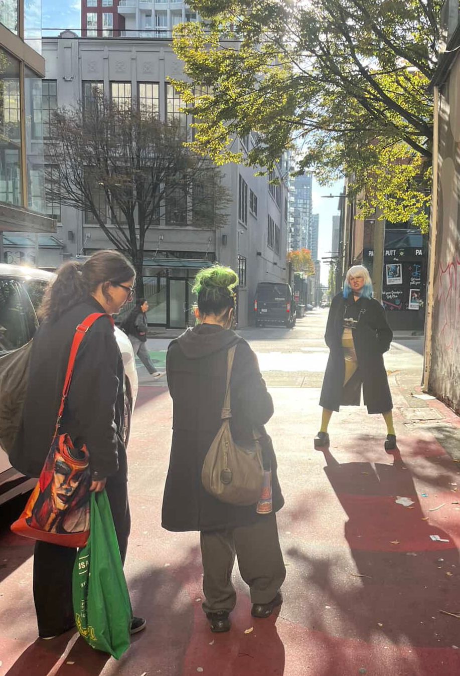 Three people stand on a sunny urban sidewalk surrounded by city buildings and autumn trees.