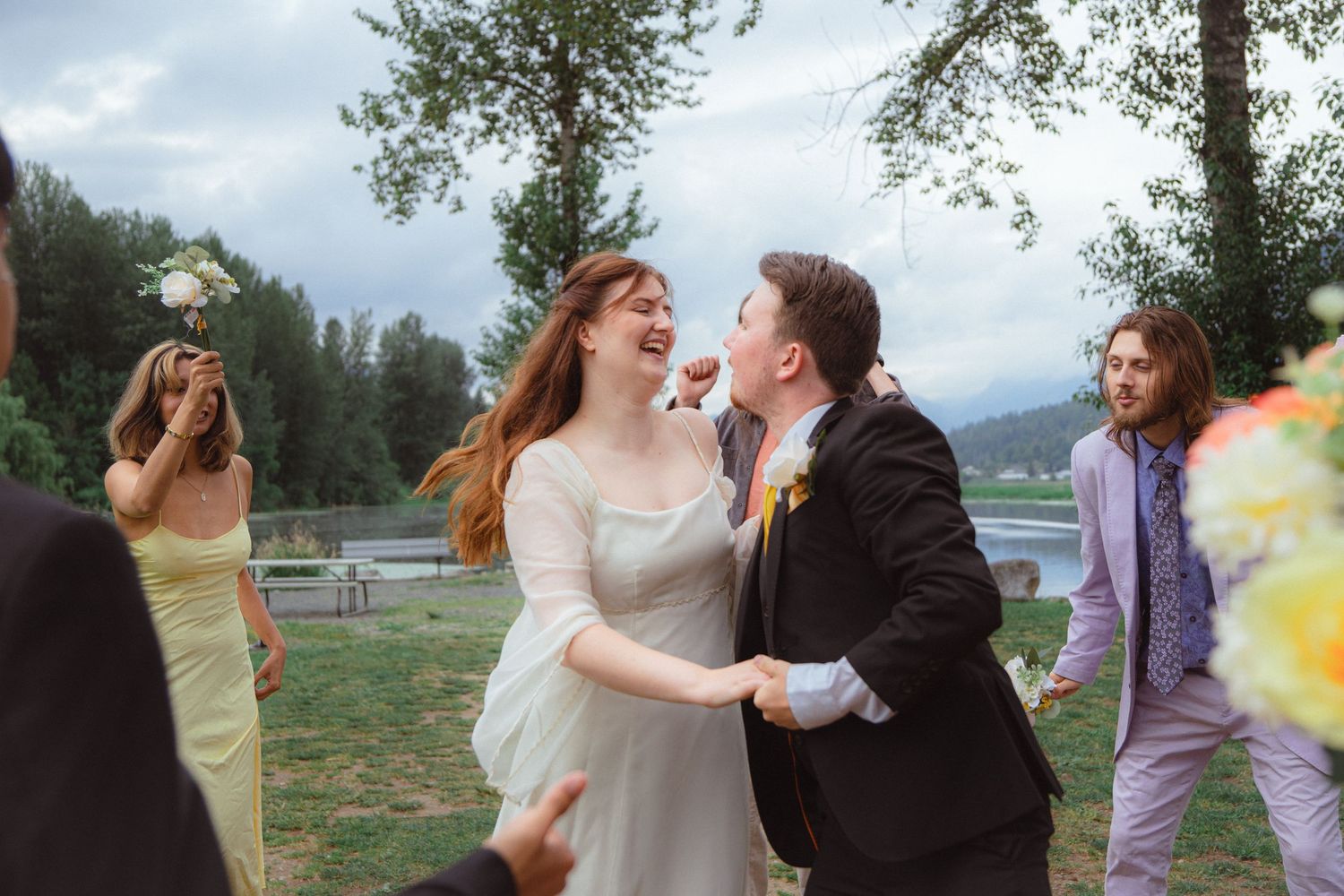 Happy newlyweds laughing and dancing together surrounded by cheering guests at outdoor wedding.