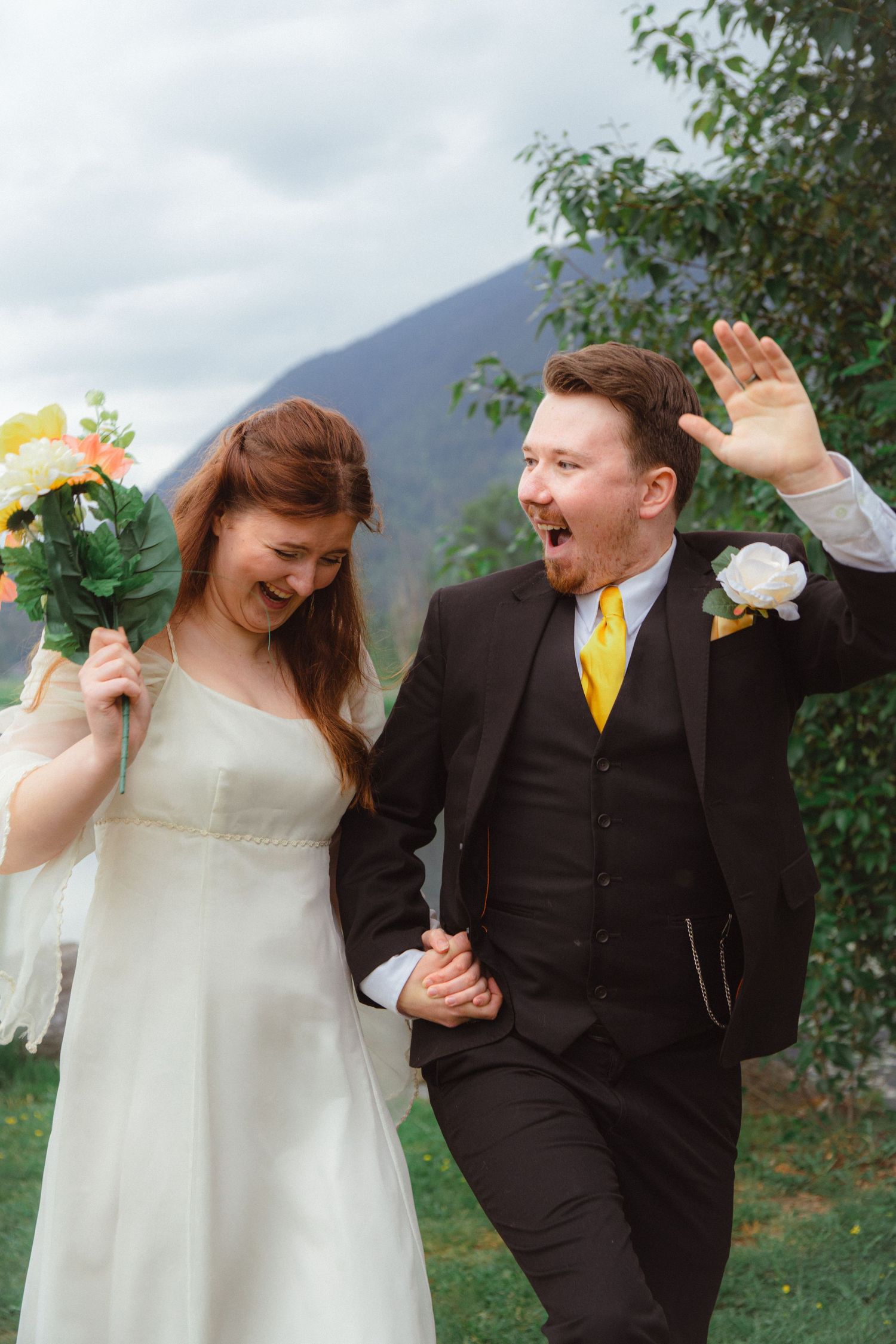 Laughing bride and groom in black suit with yellow tie celebrating outdoors with mountains behind.