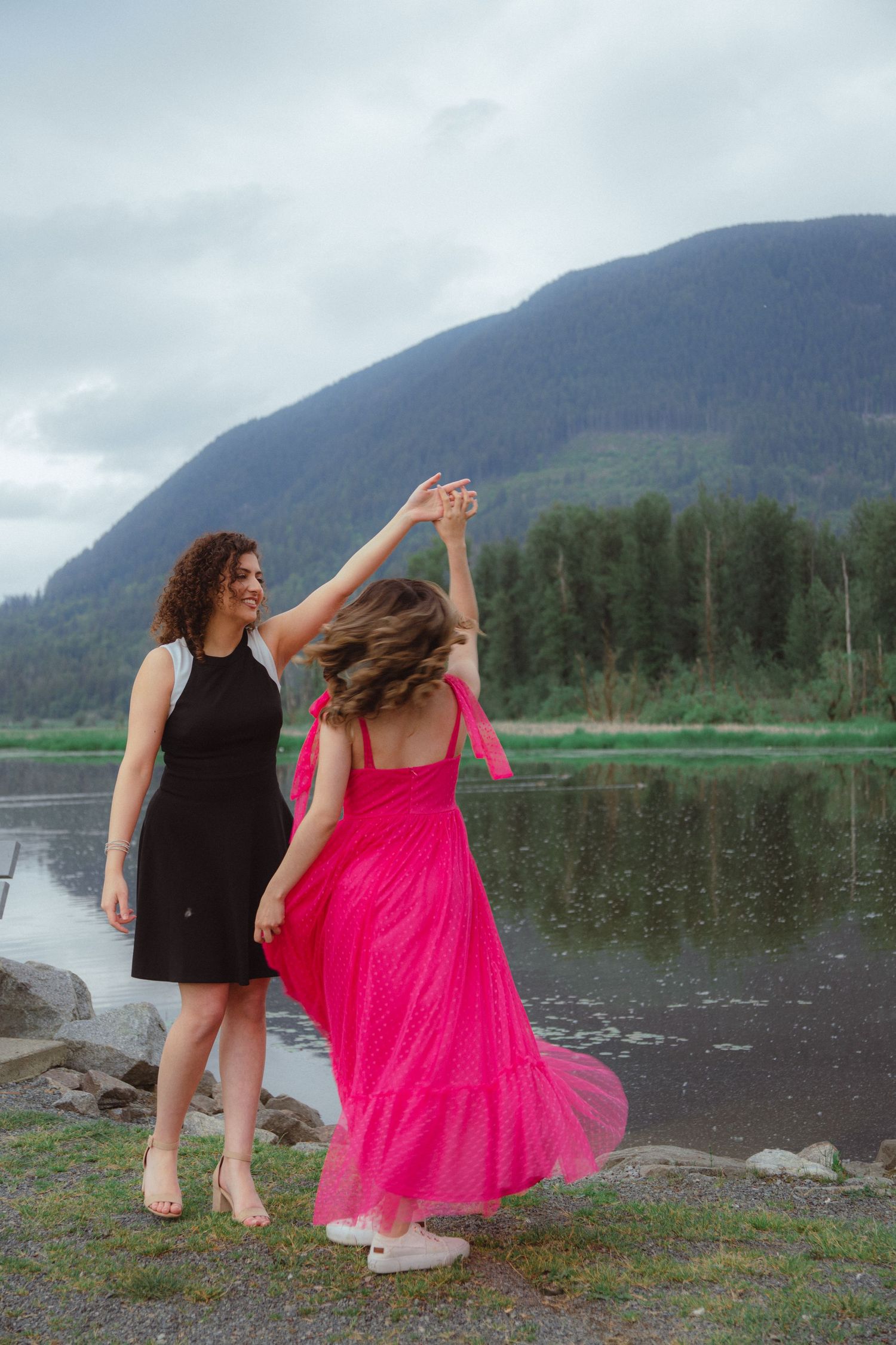 Two women dance together by a reflective mountain lake, one in a flowing pink gown.