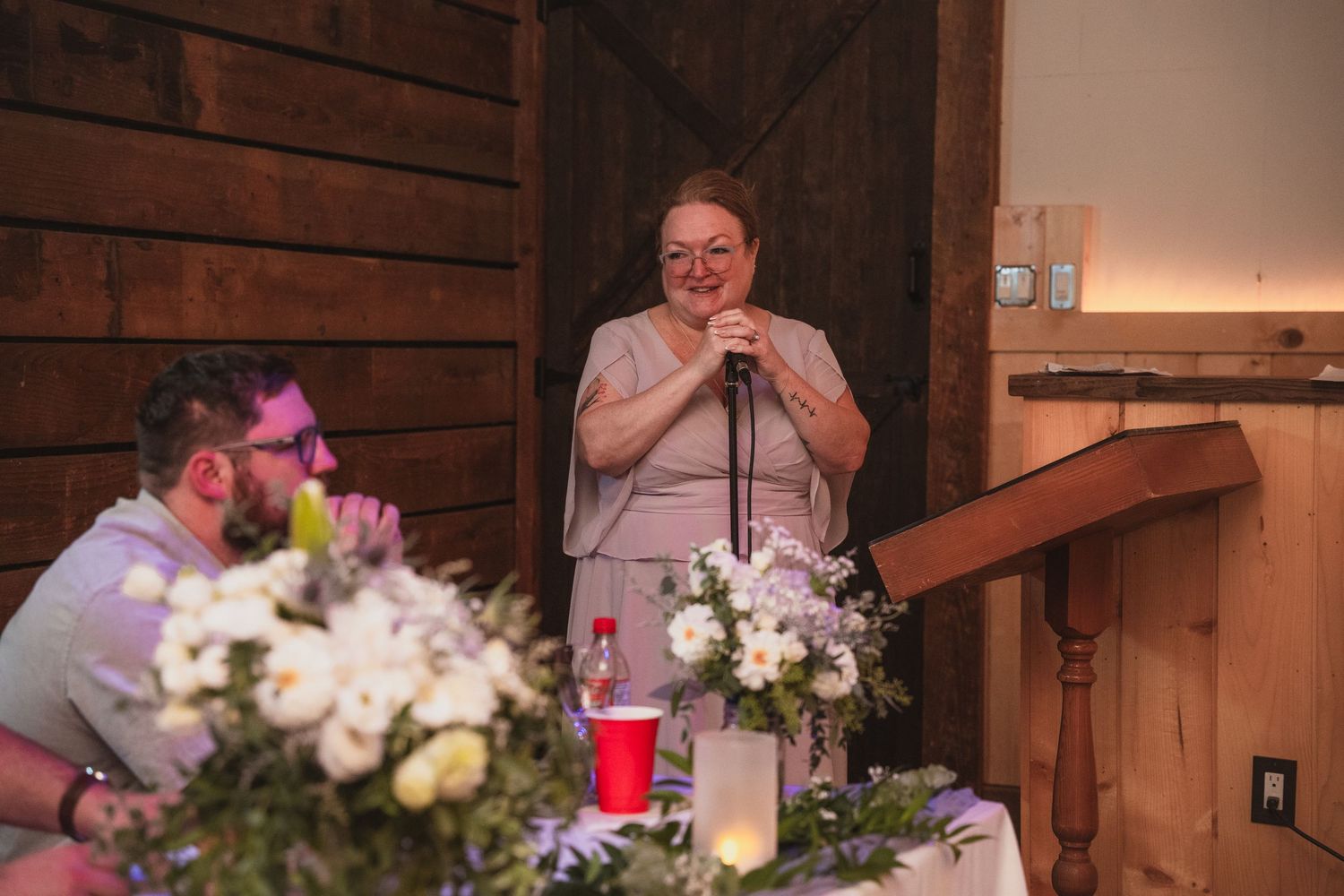 Woman delivers emotional wedding toast at microphone beside floral centerpiece in a warm barn venue.