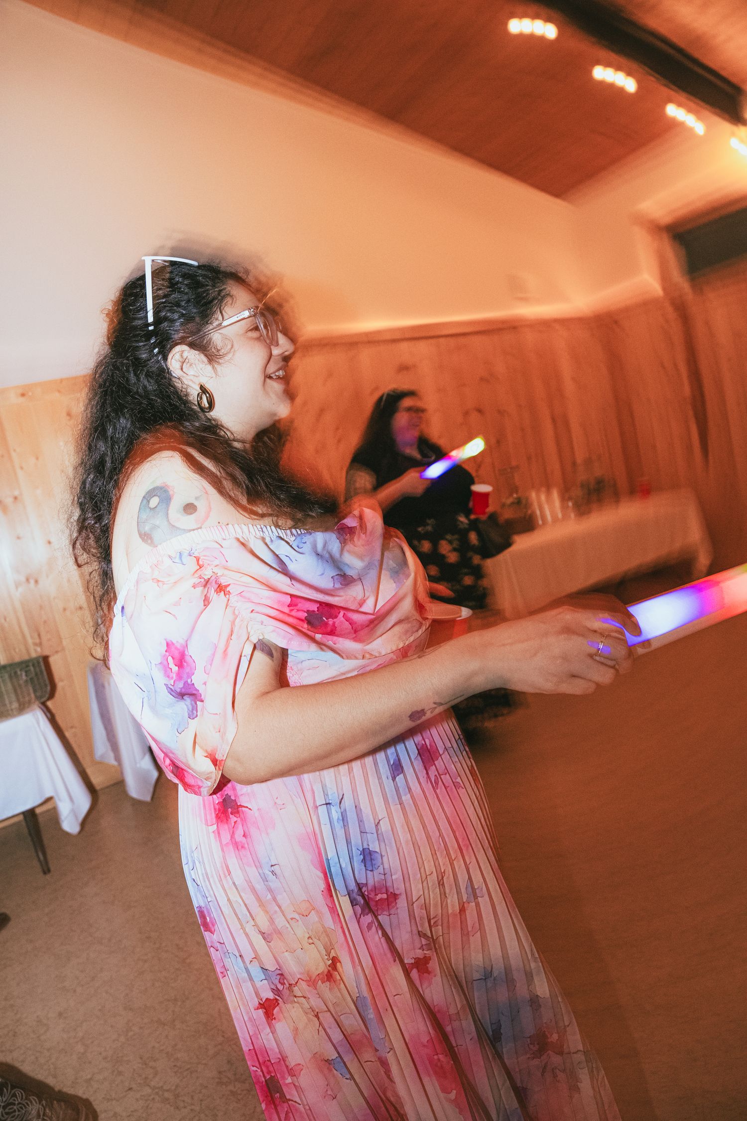Woman in colorful floral maxi dress smiling and dancing at an indoor celebration with soft warm light.