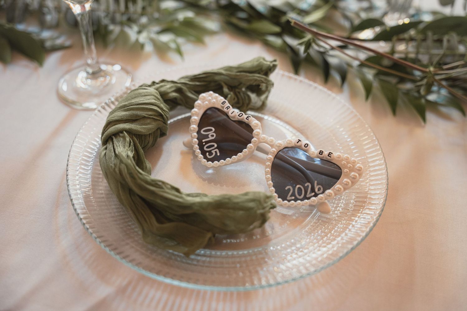 Heart-shaped sunglasses with pearl trim reading 2026 on a glass plate with sage green ribbon wedding table decor.