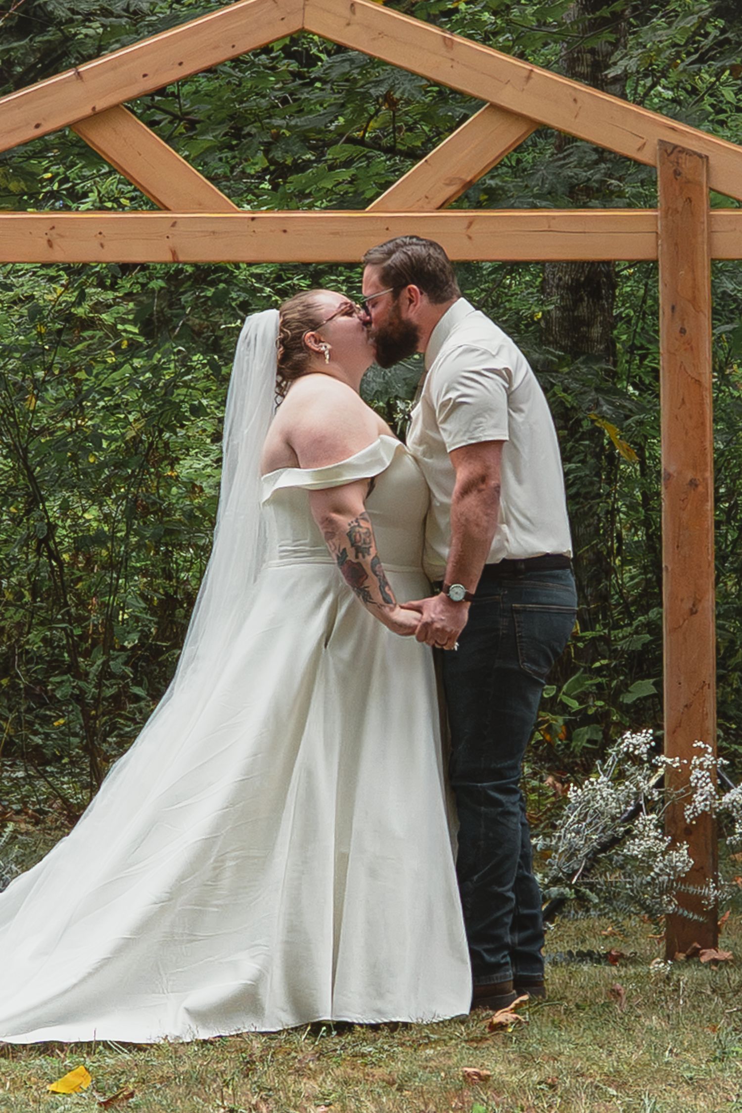 Bride and groom share first kiss under rustic wooden arch surrounded by lush green forest at outdoor wedding.