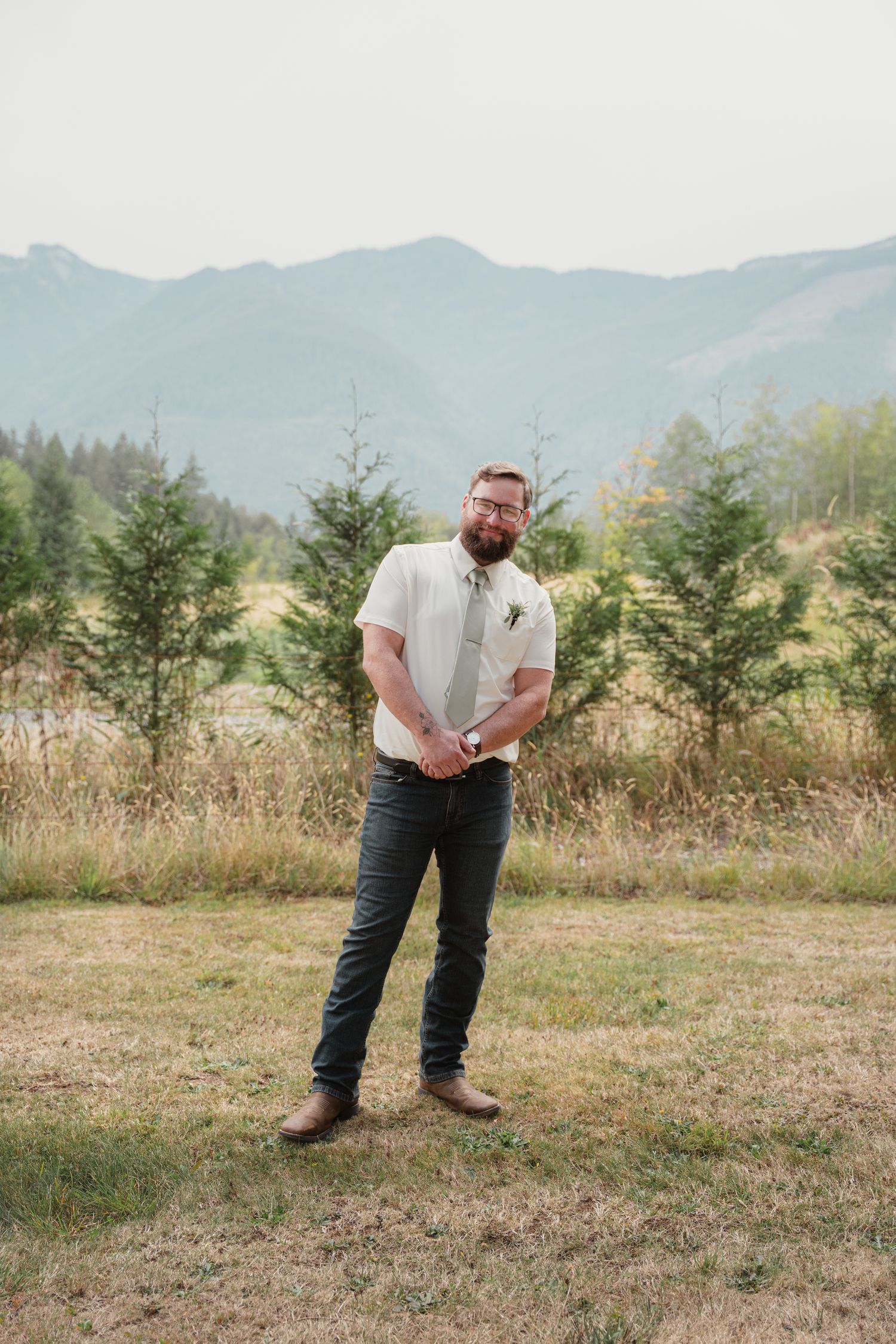 Bearded groom in white shirt and tie poses in mountain meadow at outdoor wedding.