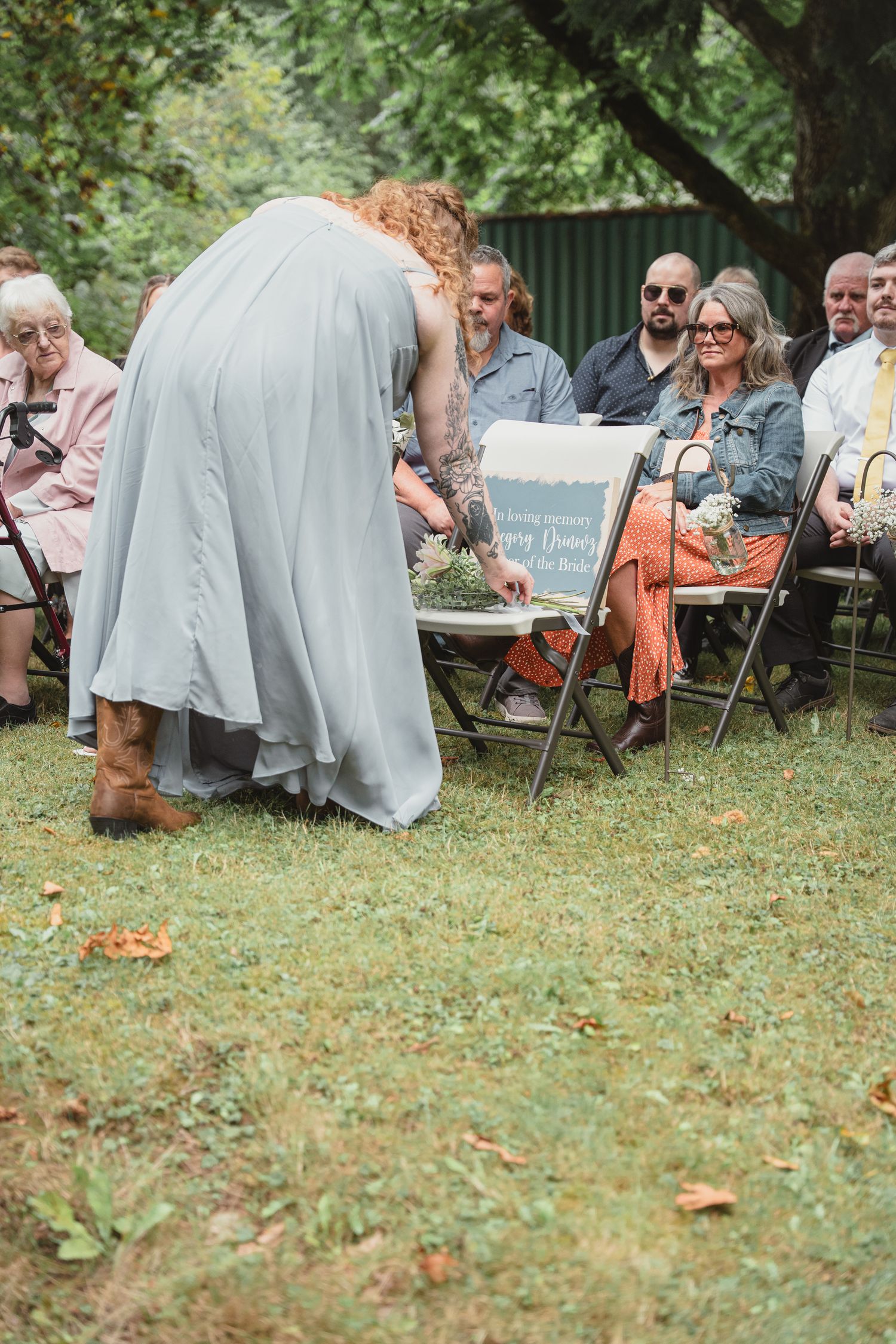 Bridesmaid in gray dress and cowboy boots leans over a memorial chair during an outdoor wedding ceremony.